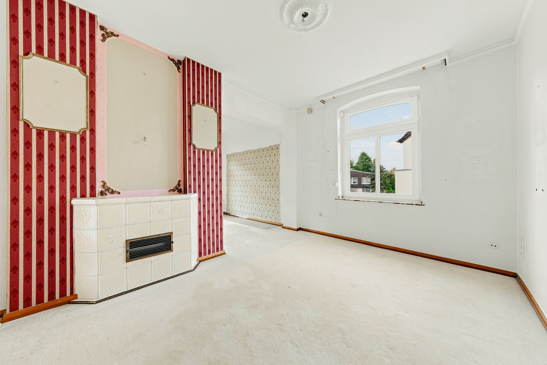 Helles Wohnzimmer mit Teppichboden, großem Fenster und Kachelofen, rote Tapete an einer Wand.