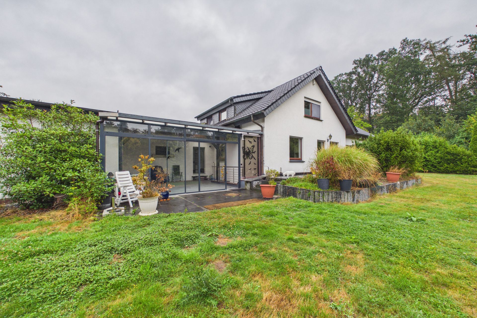 Weißes Wohnhaus mit Wintergarten und Garten mit Rasen und Pflanzen im Vordergrund.
