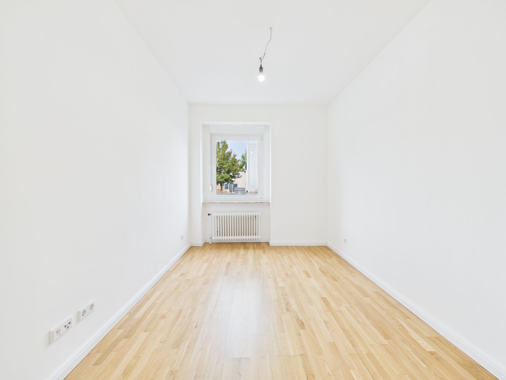 Heller, leerer Raum mit Holzfußboden, weißem Wänden und Fenster mit Heizung darunter.