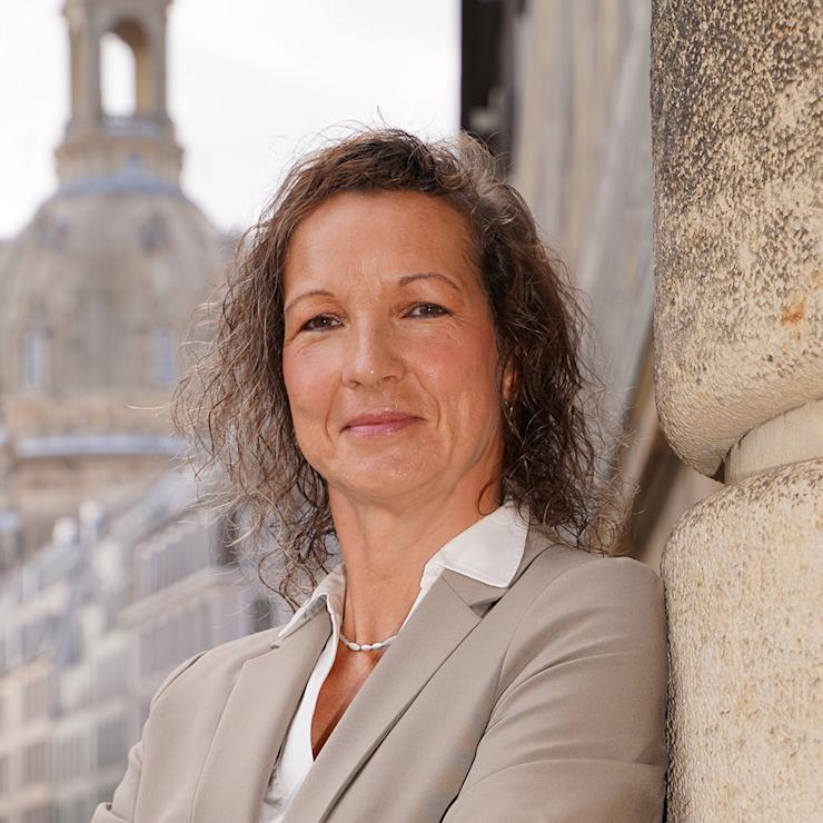 Eine Frau im beigefarbenen Anzug steht draußen vor einem historischen Gebäude.