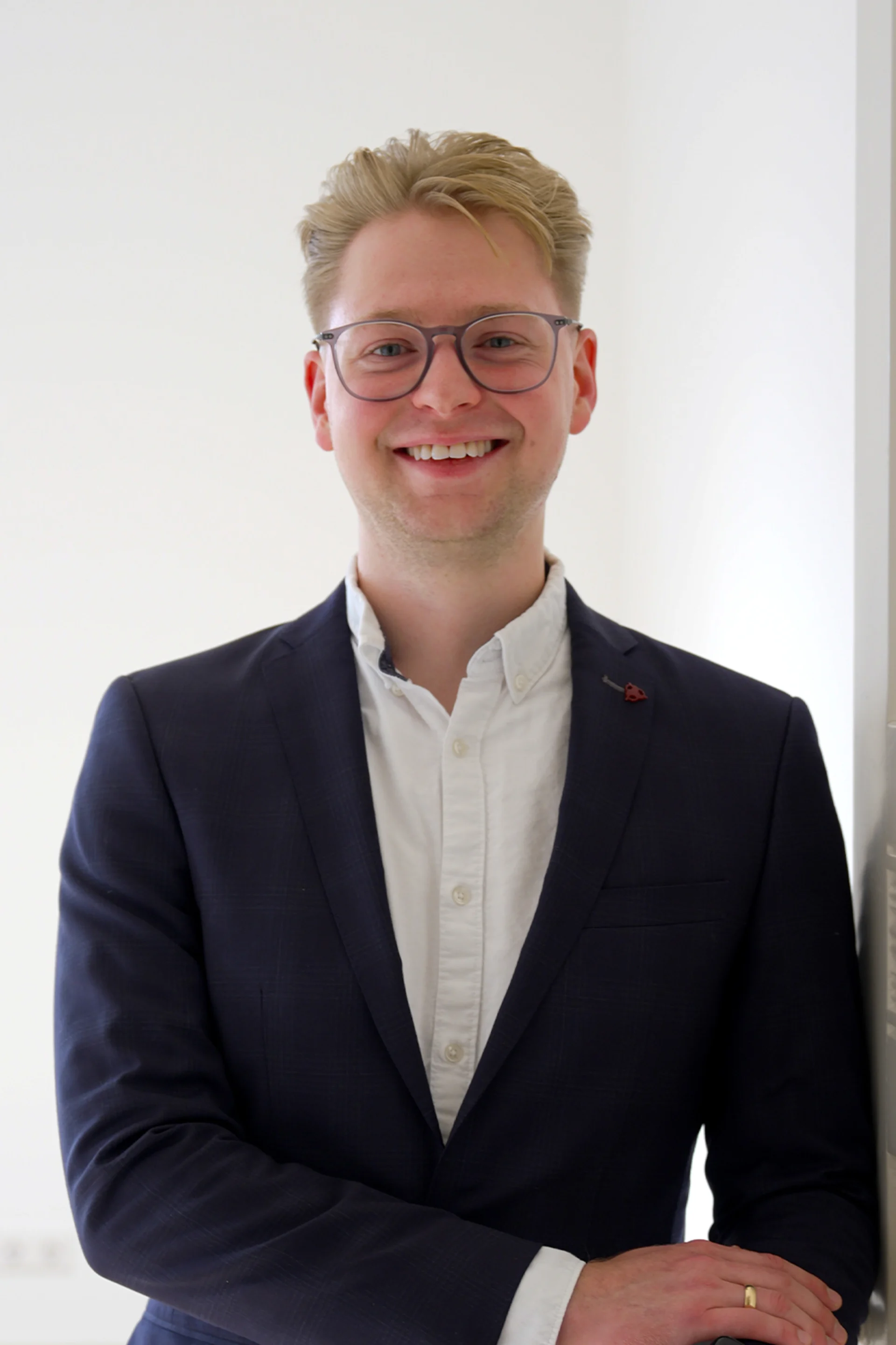 Junger Mann mit Brille und Anzug steht in einem hellen Raum und lächelt.