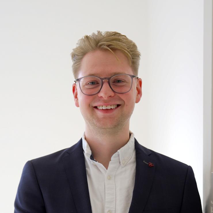 Junger Mann mit Brille und Anzug steht in einem hellen Raum und lächelt.