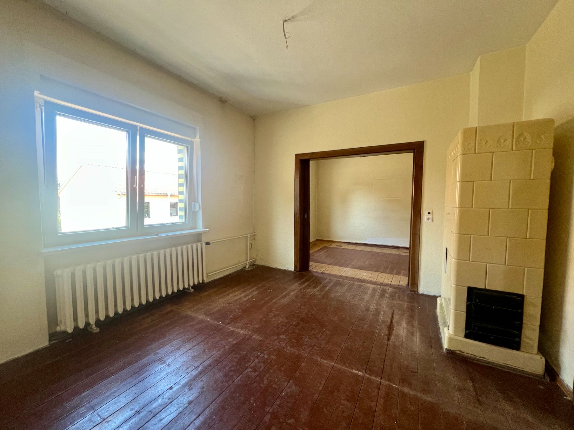 Helles Zimmer mit braunem Holzboden, Fenster, Heizkörper und Kachelofen, Zugang zu weiterem Raum.