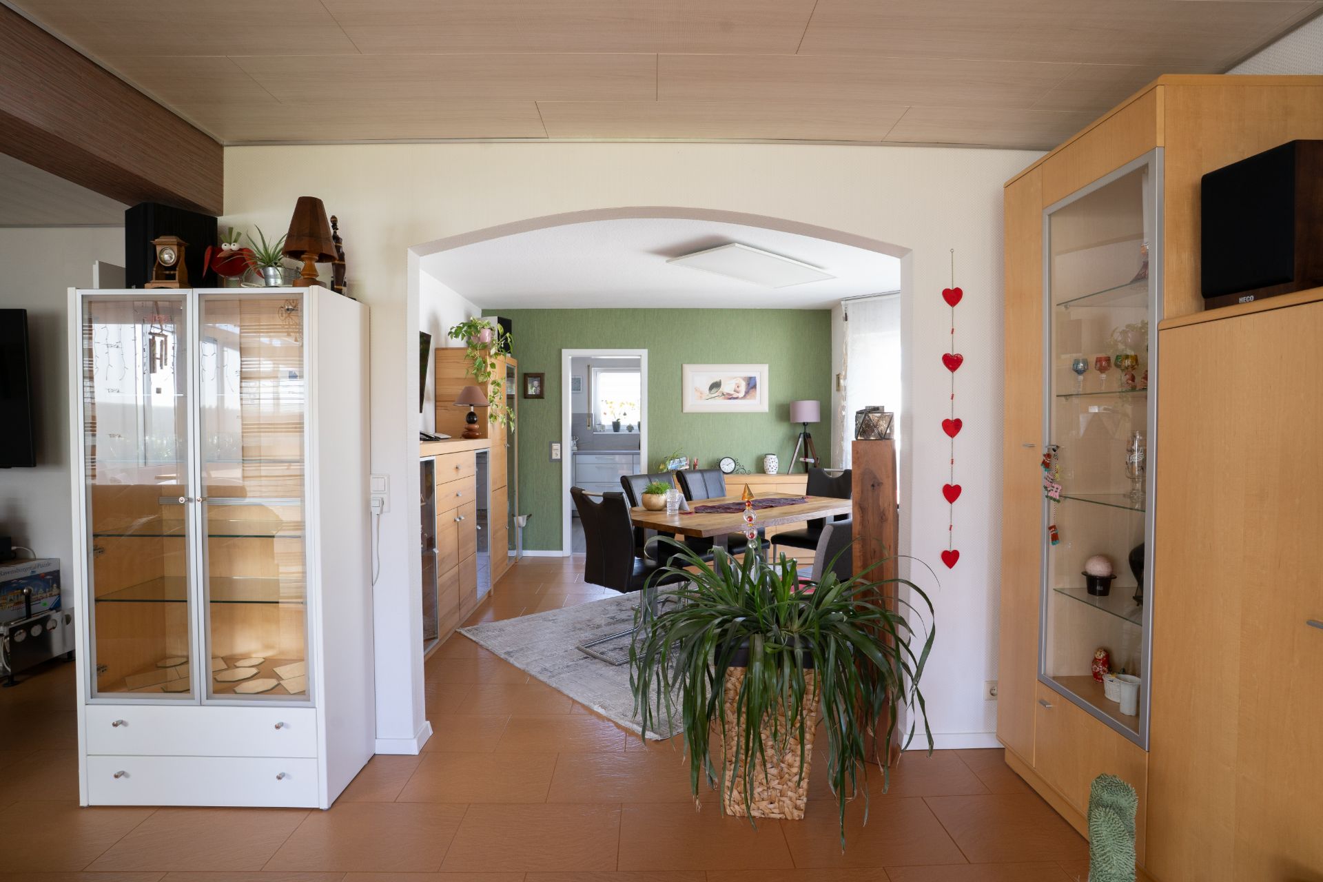 Wohnzimmer mit Vitrine, großem Zimmerpflanze und Blick auf Essbereich mit grün gestrichener Wand.
