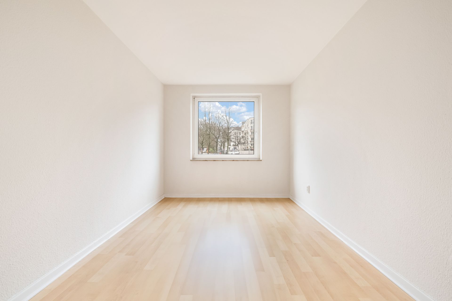 Helles, leeres Zimmer mit Holzfußboden und einem Fenster mit Blick nach draußen.