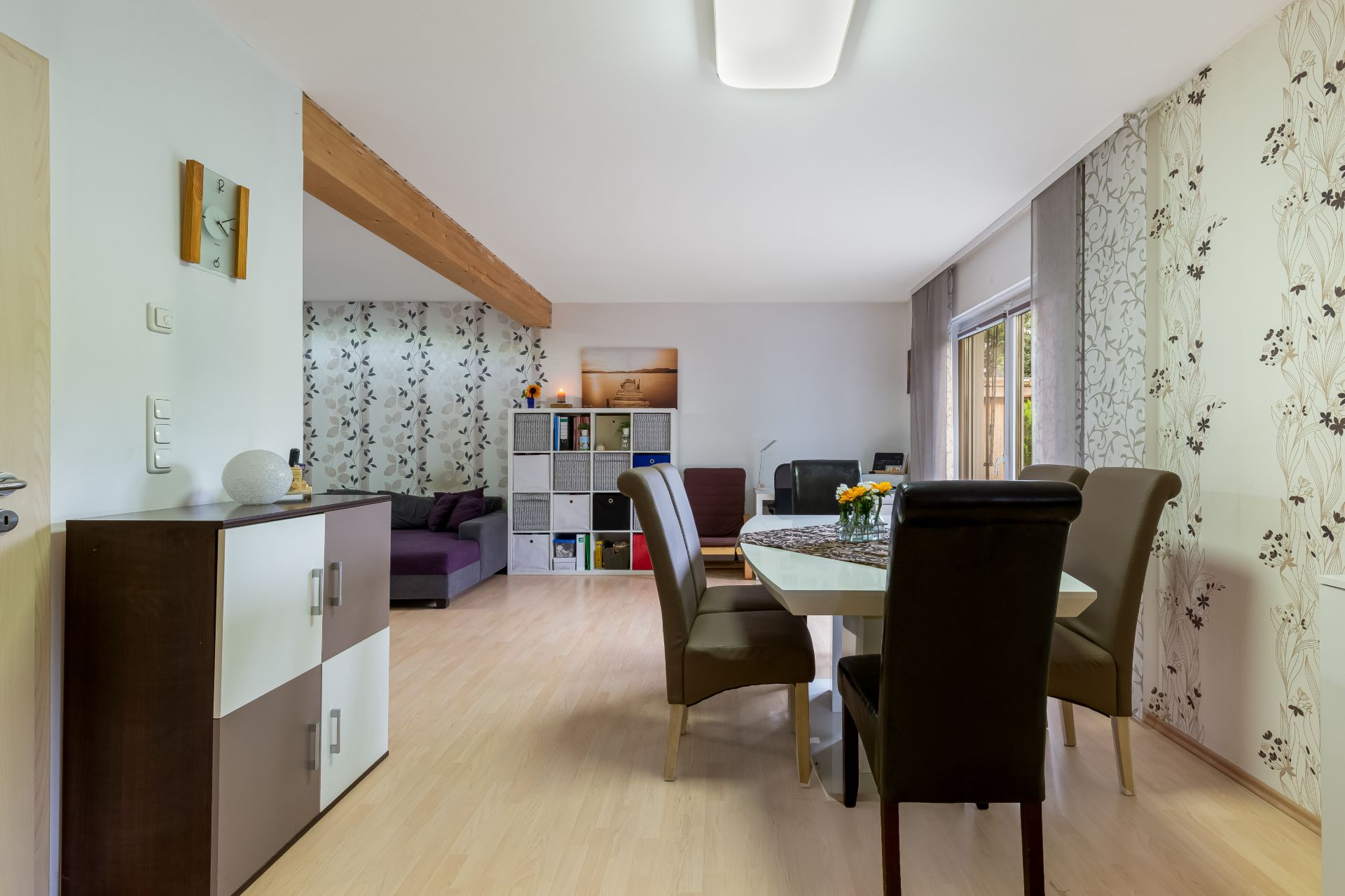 Helles Wohnzimmer mit Esstisch, sechs Stühlen, Regalwand und Sofa, Holzboden und floral gemusterter Tapete.