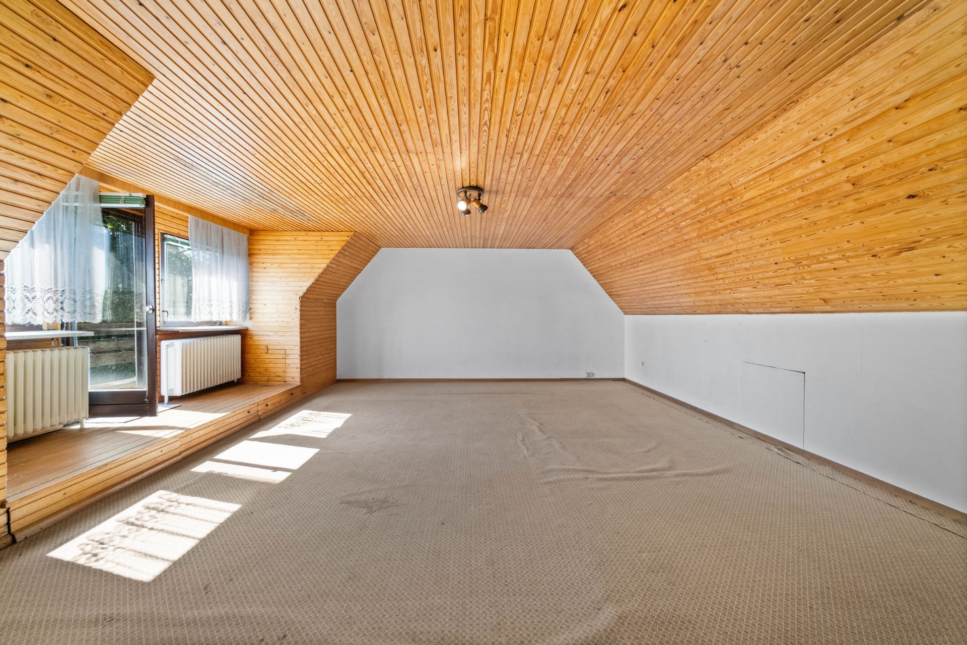 Leeres Dachzimmer mit Holzdecke, Teppichboden und Fenstern mit Vorhängen.