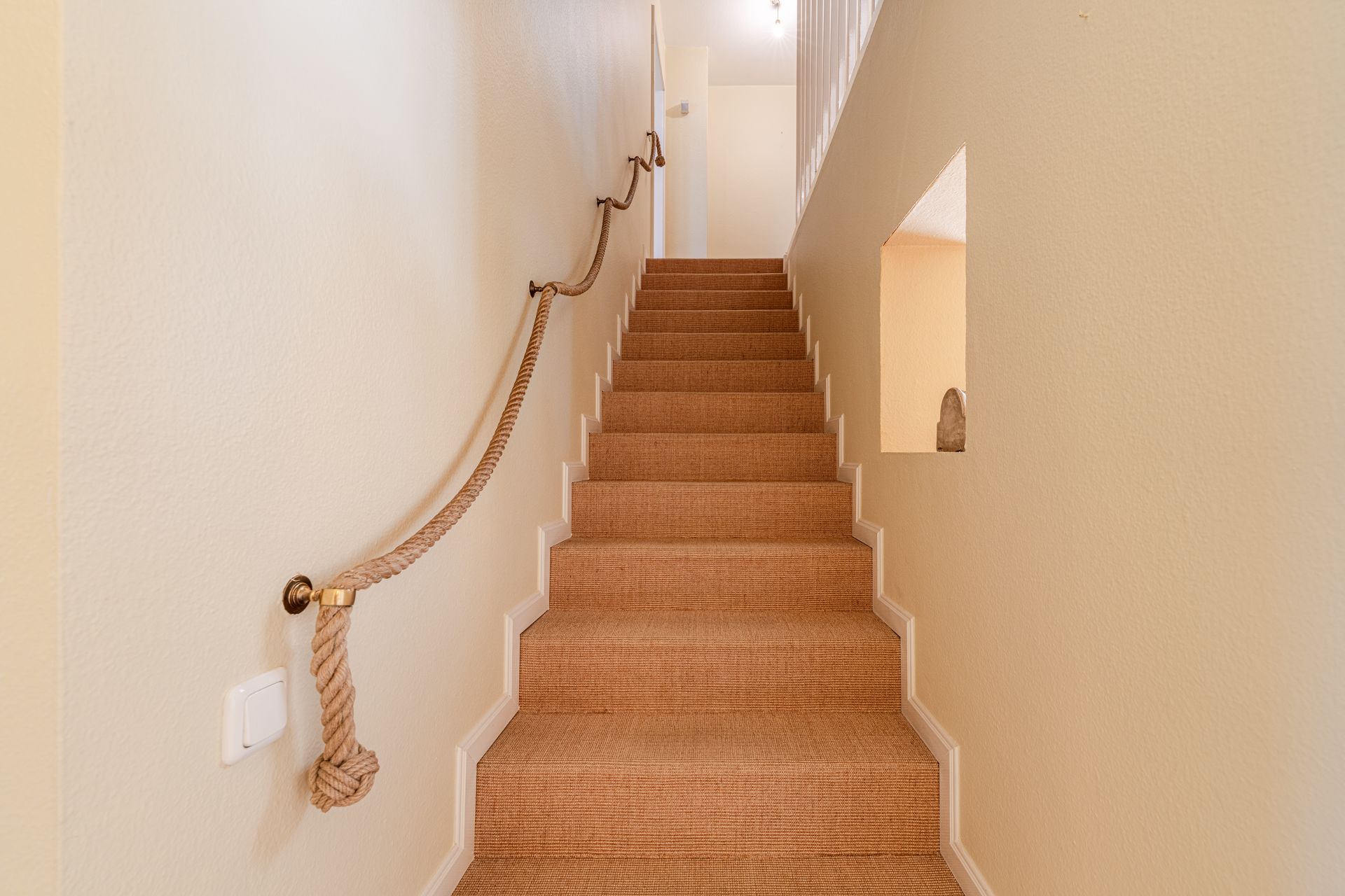 Teppichbezogene Treppe mit Seil als Handlauf in einem Flur mit cremefarbenen Wänden.