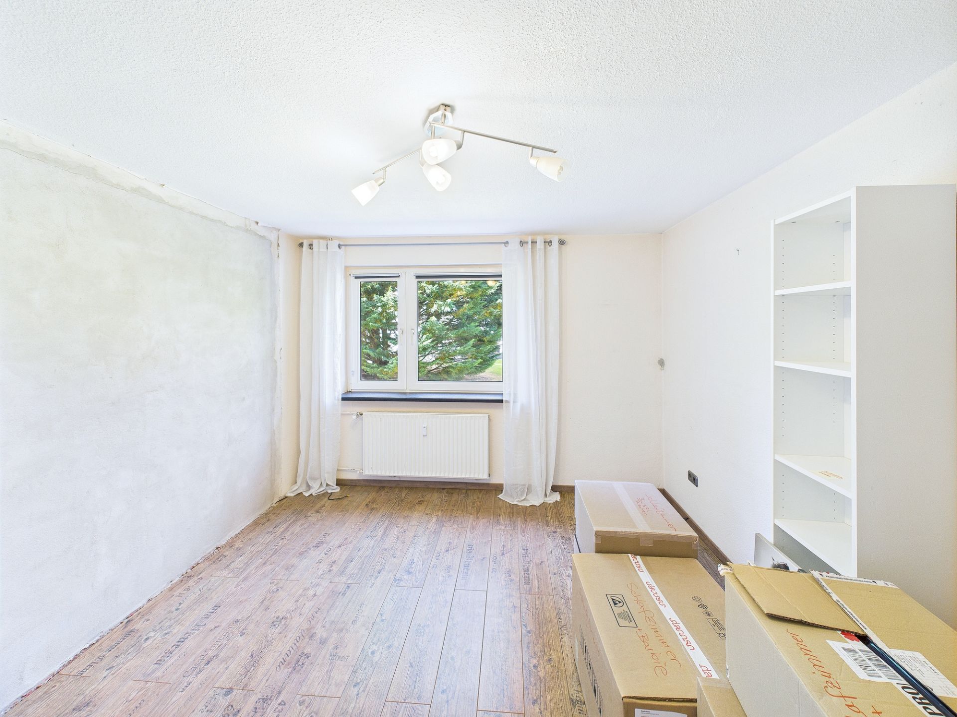 Helles Zimmer mit Fenster, Vorhängen, Regal und Umzugskartons auf Holzfußboden.