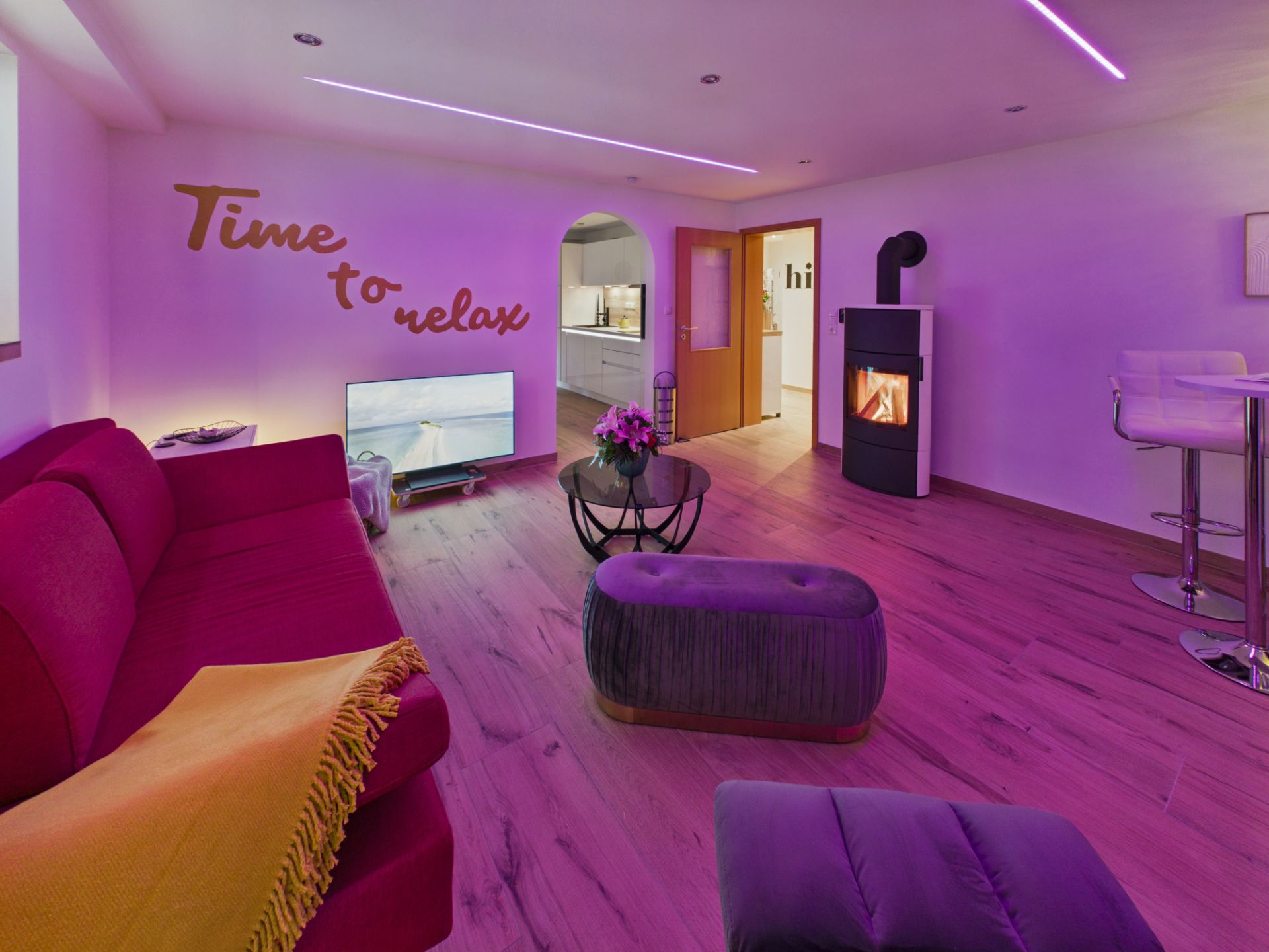 Modernes Wohnzimmer mit lila Beleuchtung, Sofa, Fernseher, Kamin und offener Küche.
