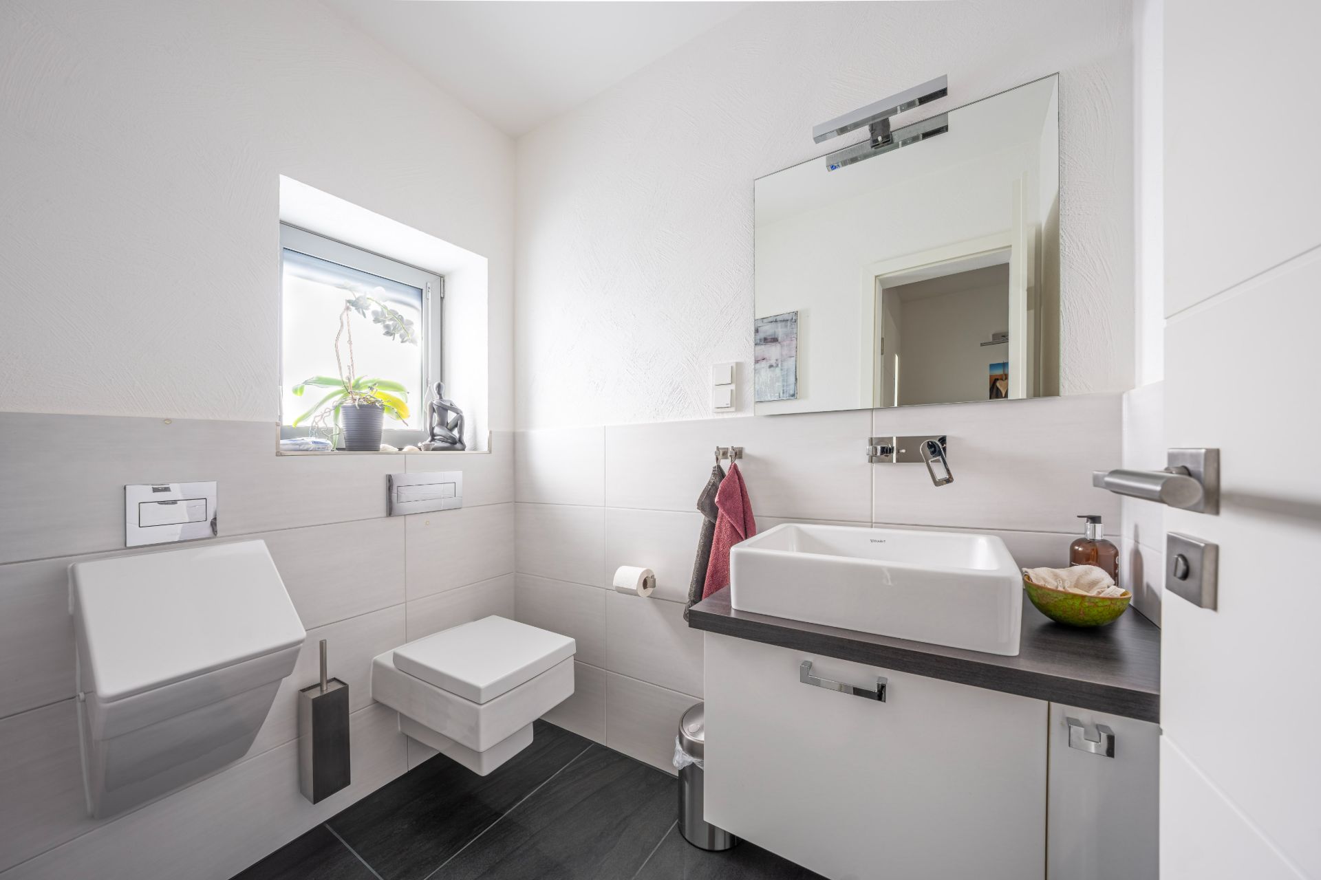 Modernes Badezimmer mit Wandtoilette, Urinal, Waschbecken und kleinem Fenster mit Pflanze.