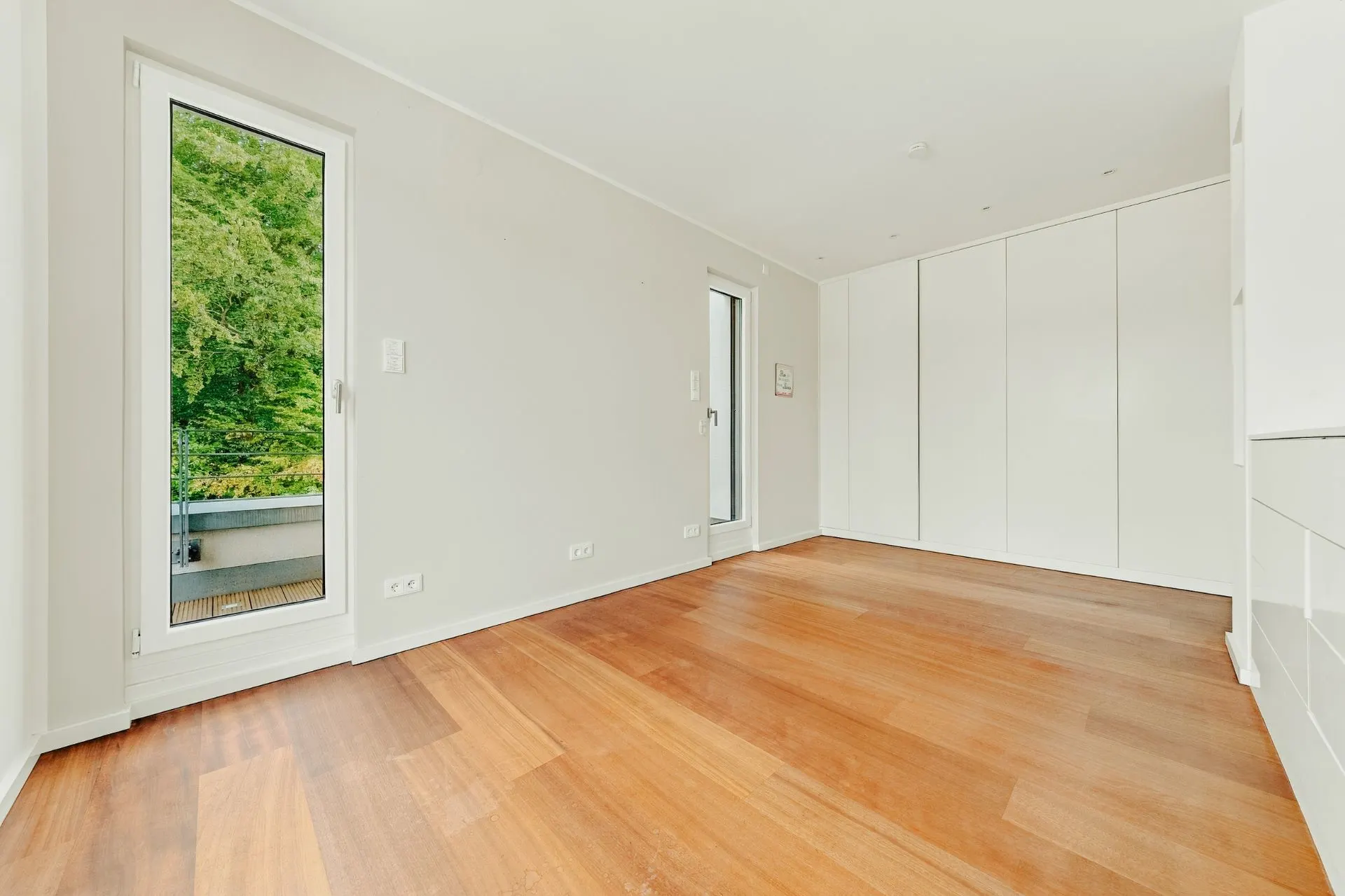 Helles Zimmer mit Holzboden, weißen Wänden, zwei Fenstertüren und Einbauschränken.