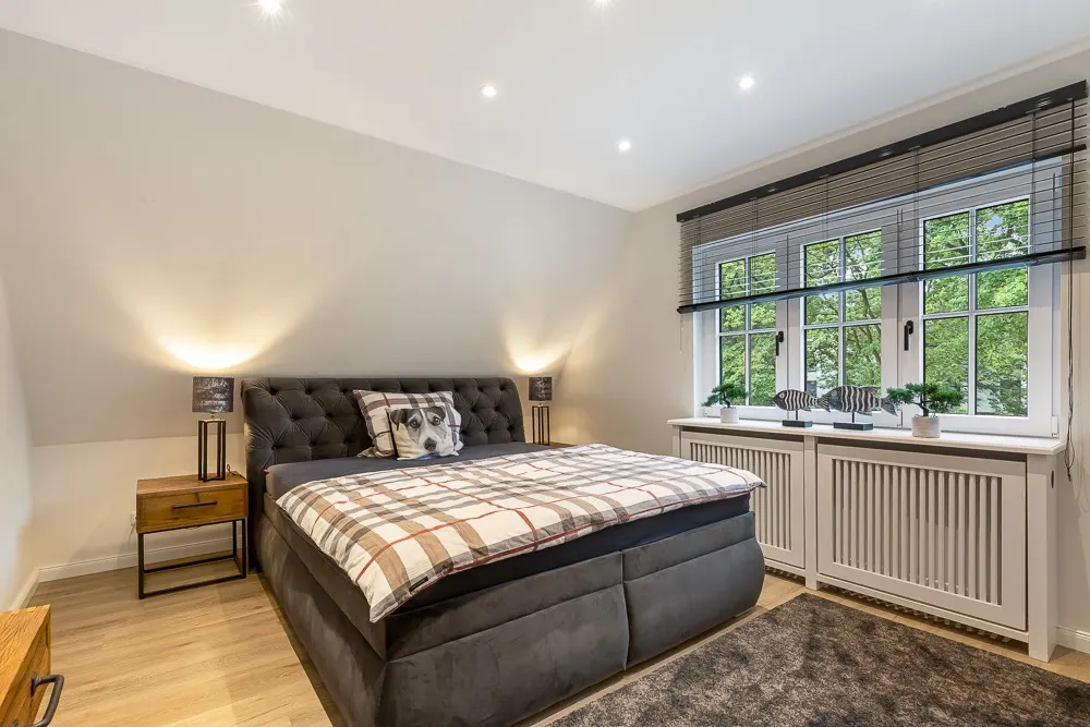 Schlafzimmer mit Doppelbett, Nachttischen, Fenster mit Jalousien und Pflanzen auf der Fensterbank.