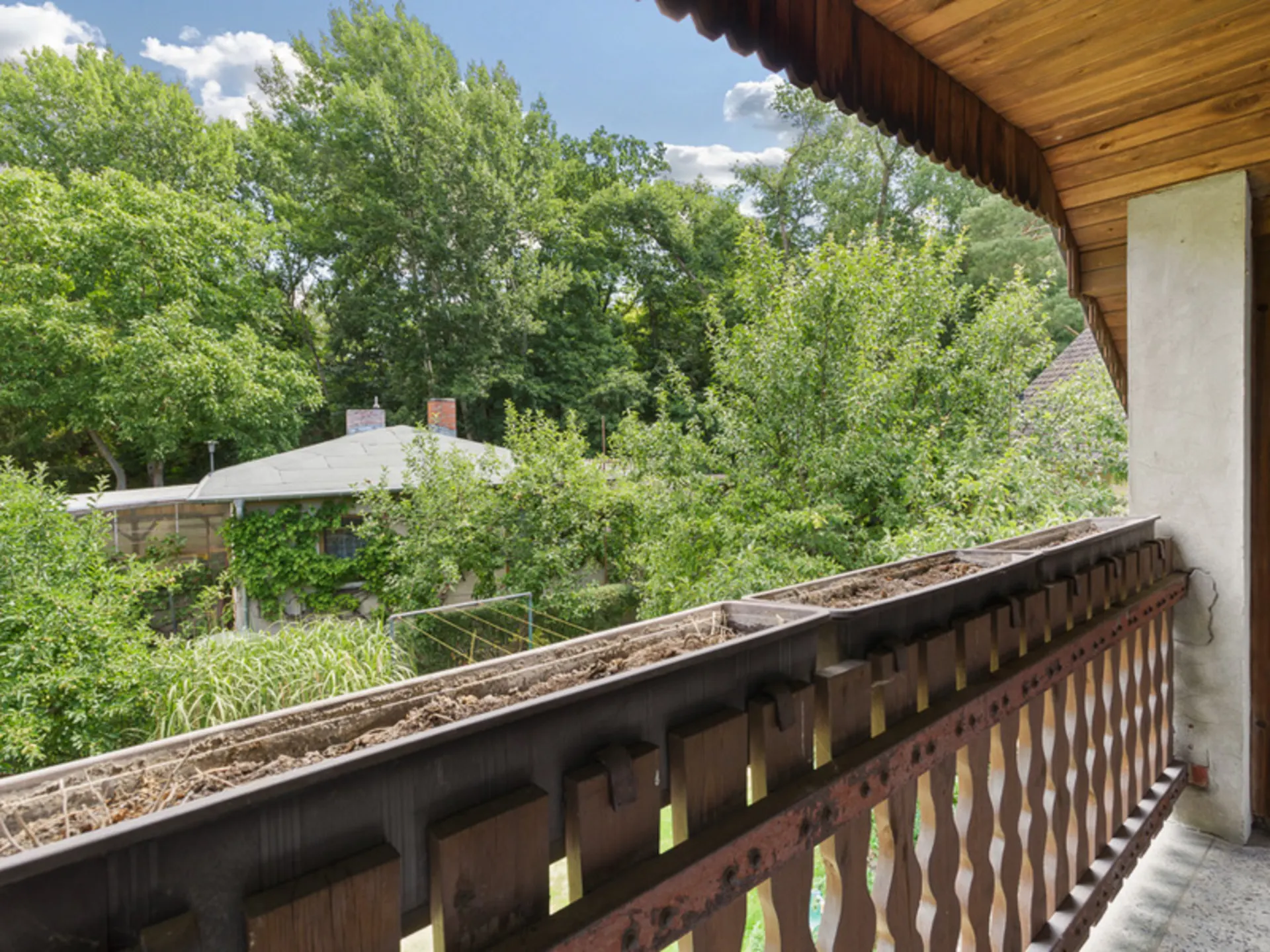 Holzbalkon mit Blumenkasten und Blick auf grüne Bäume und ein Hausdach.