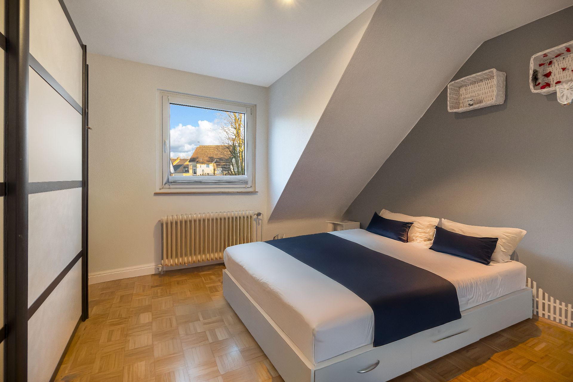 Schlafzimmer mit Doppelbett, Kleiderschrank und Fenster, darunter eine Heizung.