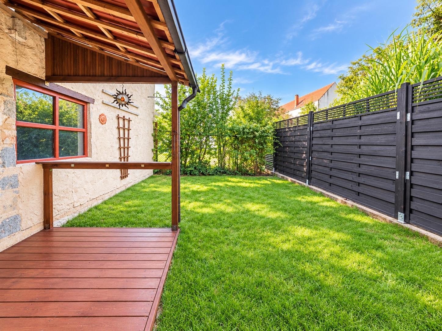 Kleiner Garten mit Holzzaun, Rasenfläche und überdachter Holzterrasse neben einem Haus.