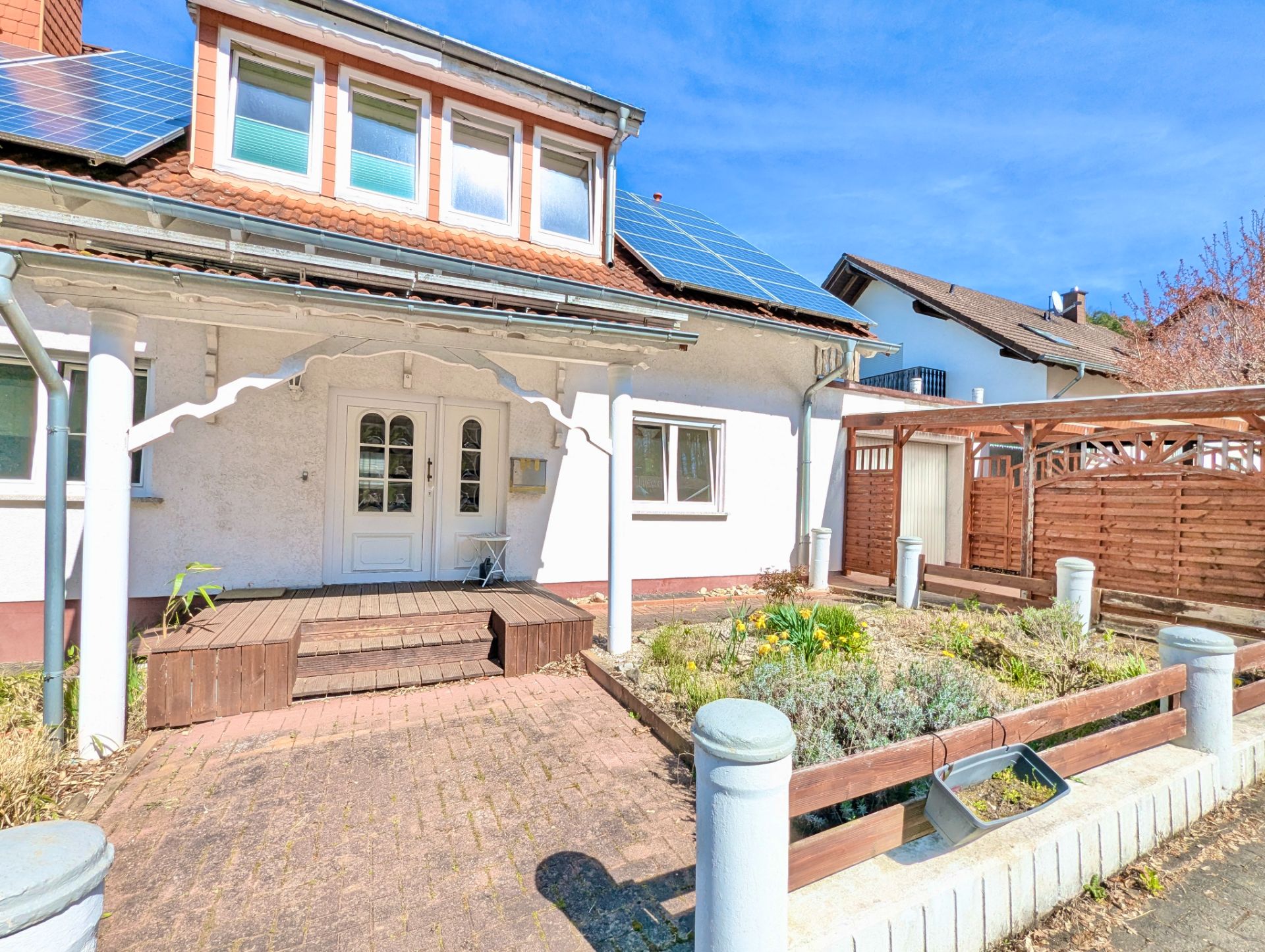 Vorderansicht eines Einfamilienhauses mit Vorgarten, Holzterrasse und Solarpanels auf dem Dach.