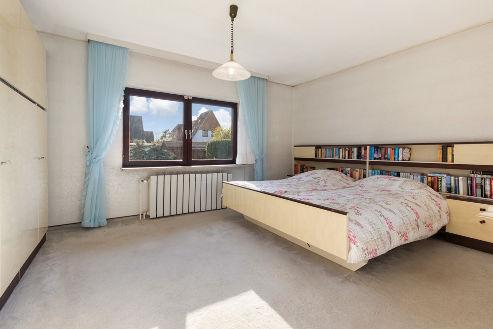 Helles Schlafzimmer mit Doppelbett, Bücherregal im Kopfteil und Fenster mit blauen Vorhängen.
