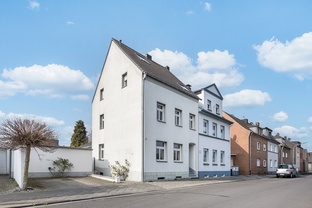 Weißes Wohnhaus mit Satteldach an einer ruhigen Straße, neben anderen Häusern.