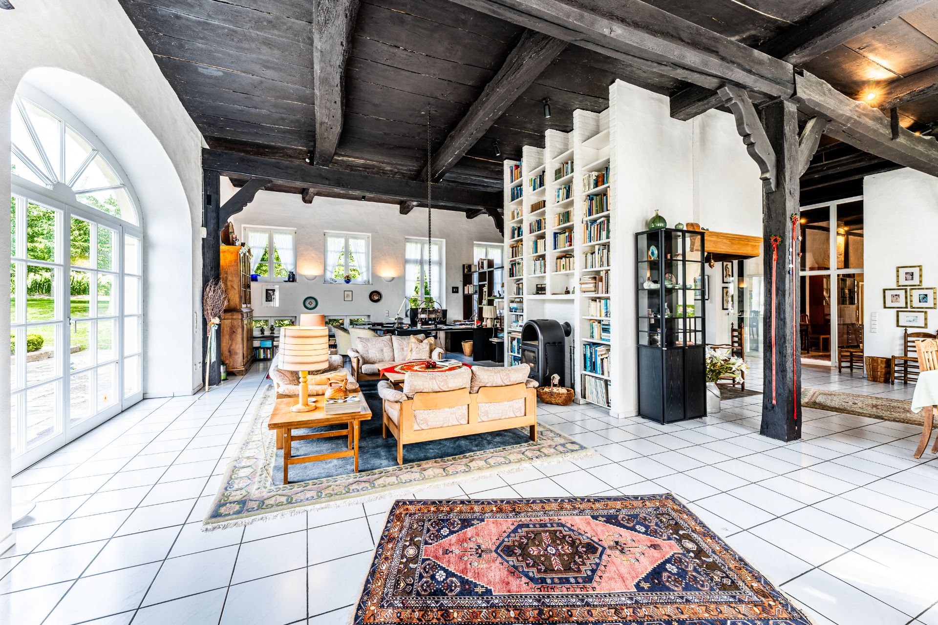 Heller Wohnraum mit großen Fenstern, Holzbalkendecke, Sofa, Bücherregal und Teppichen.