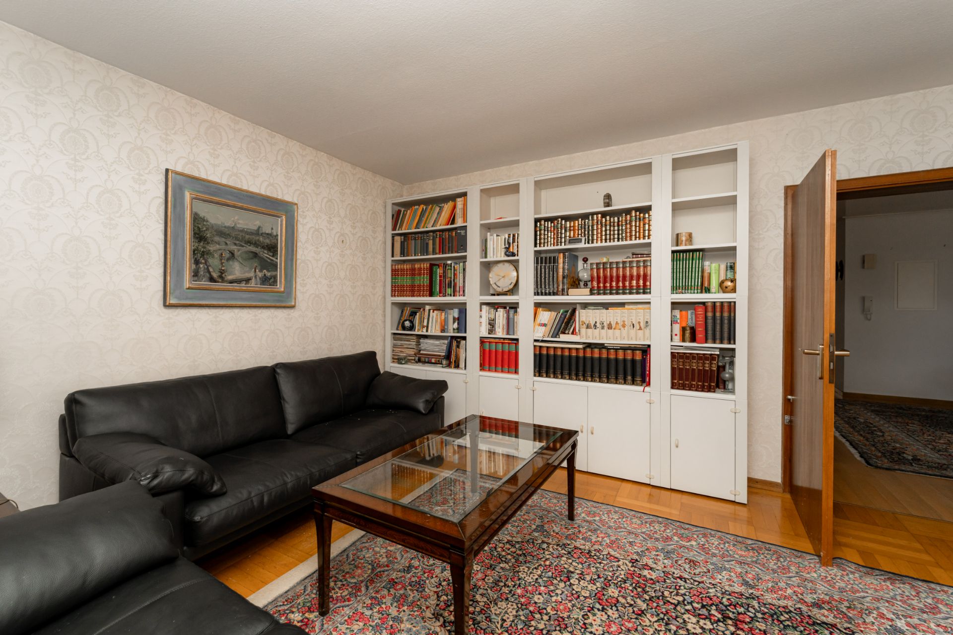 Wohnzimmer mit schwarzen Ledersofas, Bücherregal und Glastisch auf Teppich.