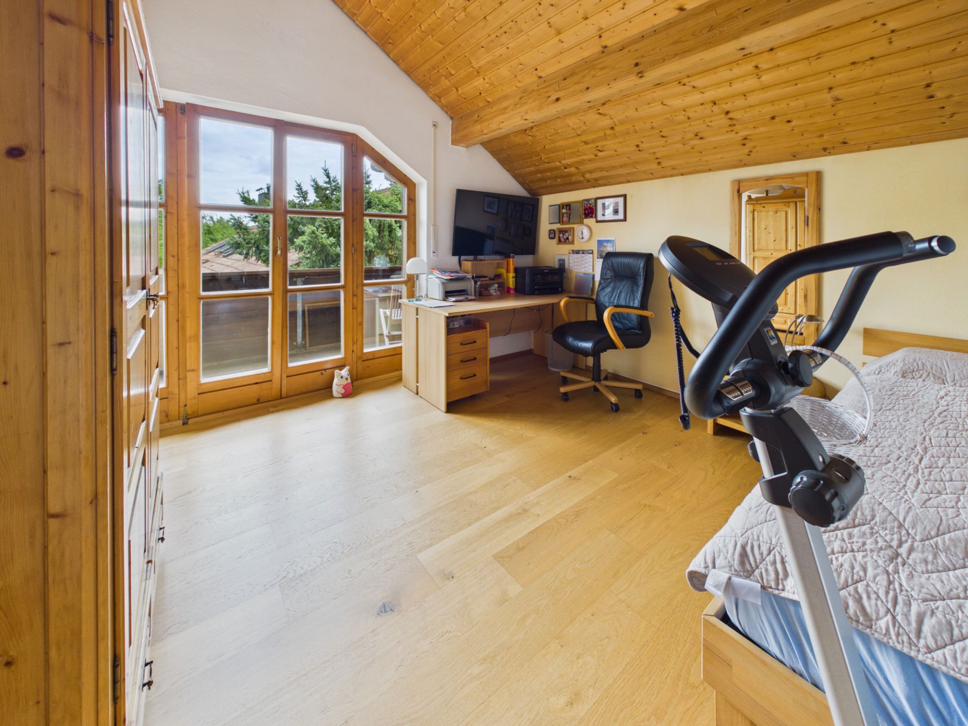 Helles Zimmer mit Holzdecke, Schreibtisch, Stuhl, Bett und Heimtrainer, große Fenster und Balkon.