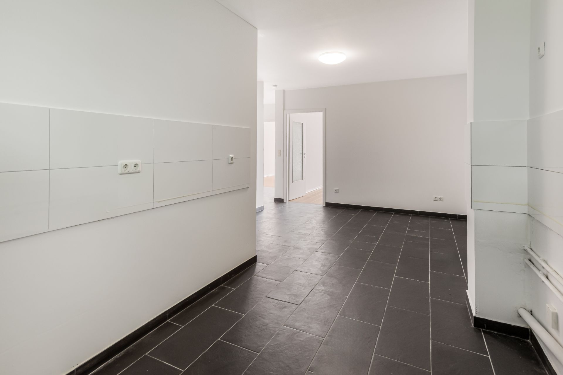 Leerer Raum mit dunklem Fliesenboden, weißen Wänden und sichtbaren Steckdosen.