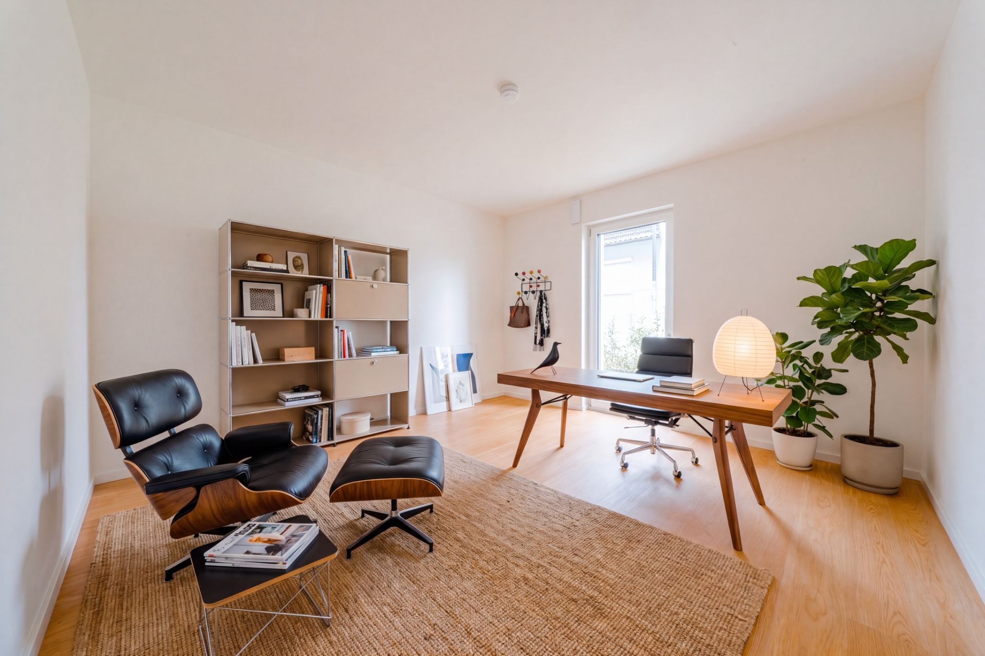 Helles Arbeitszimmer mit Schreibtisch, Regal, Sessel, Pflanze und großem Fenster.