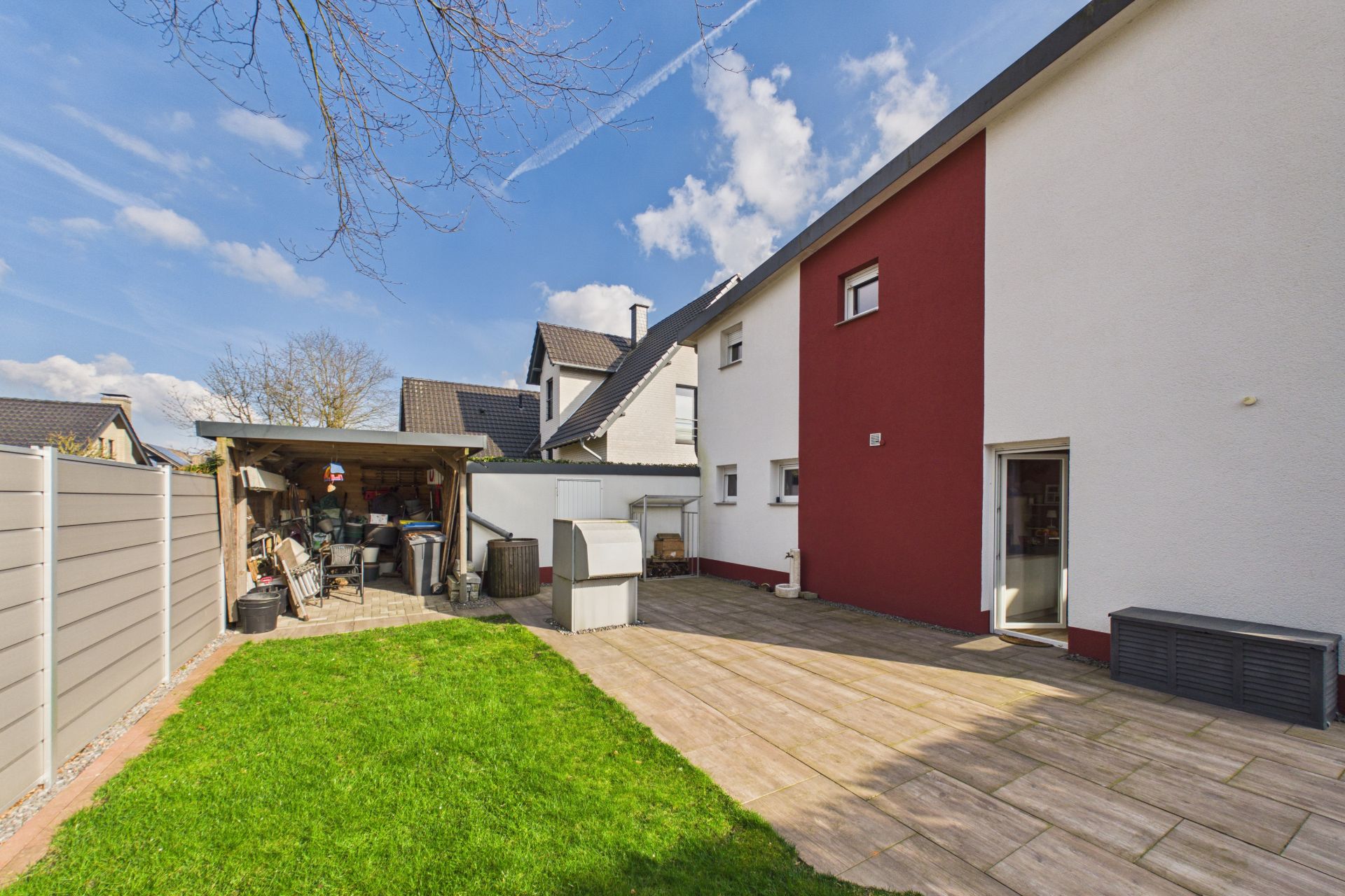 Haus mit weiß-roter Fassade, Terrasse mit Fliesen und überdachtem Abstellbereich im Garten.