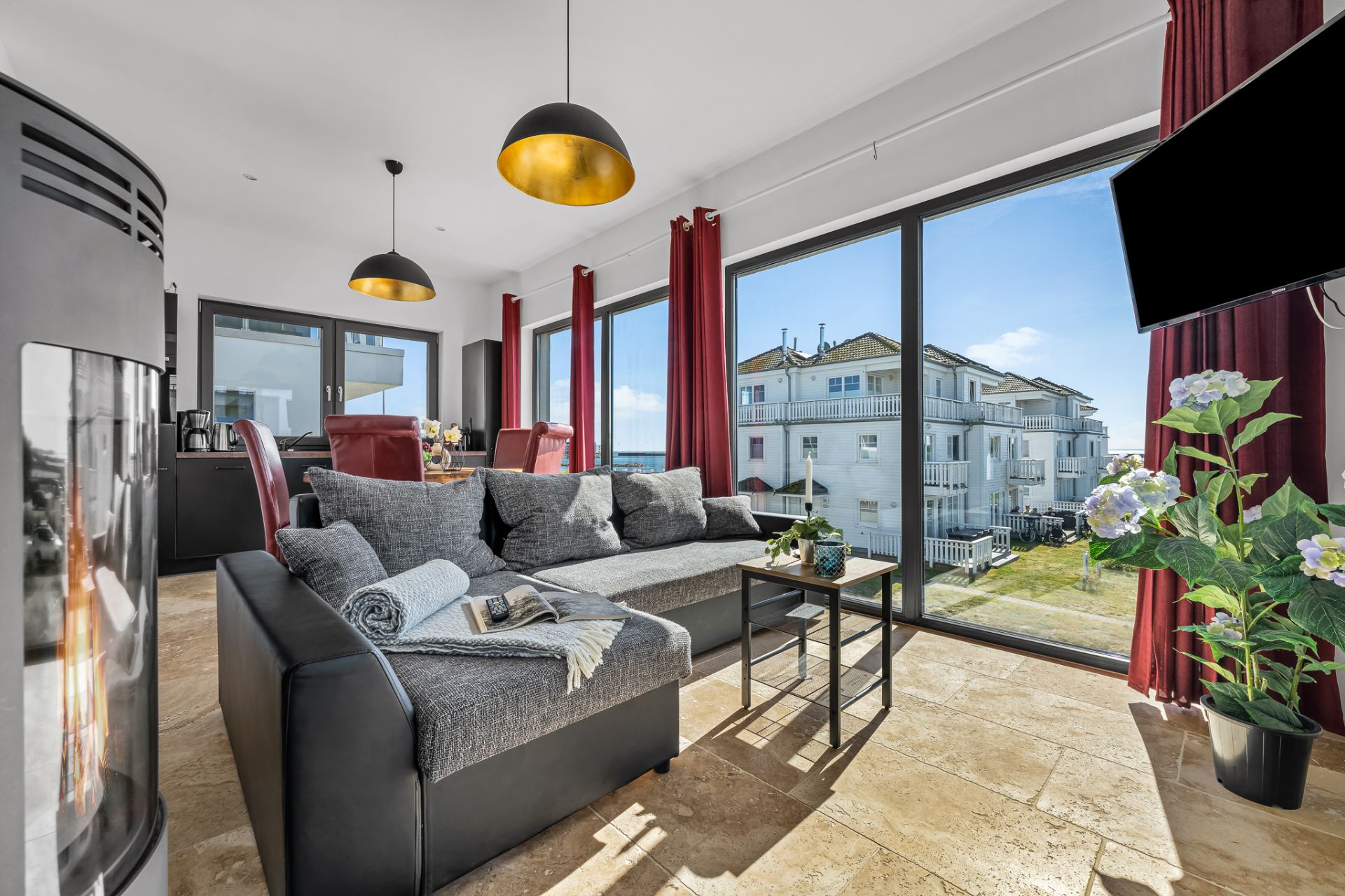Helles Wohnzimmer mit grauem Sofa, Kamin, großen Fenstern und Blick auf ein Gebäude draußen.