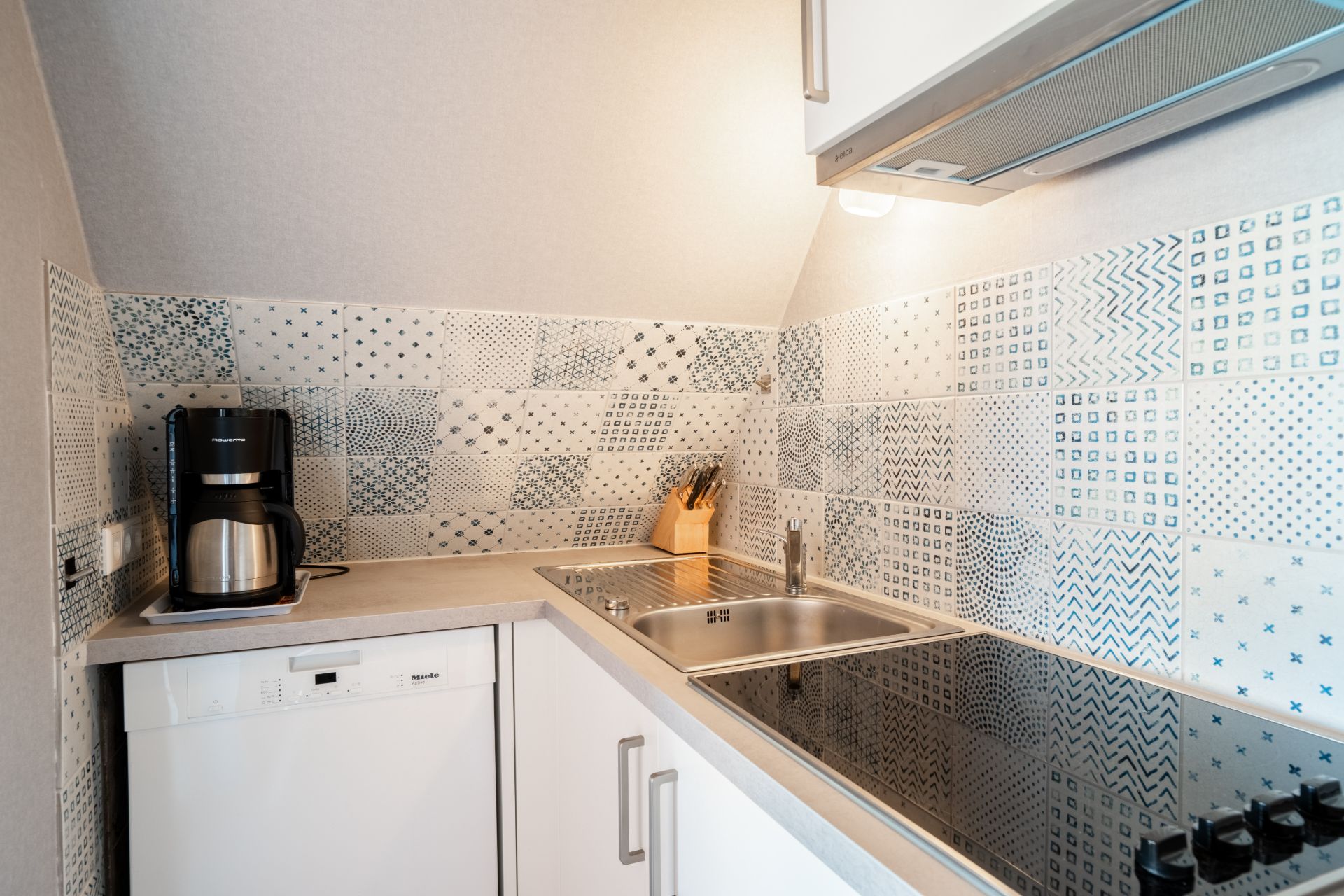 Kleine moderne Küche mit Kaffeemaschine, Spüle, Kochfeld und gemusterten Fliesen an der Wand.