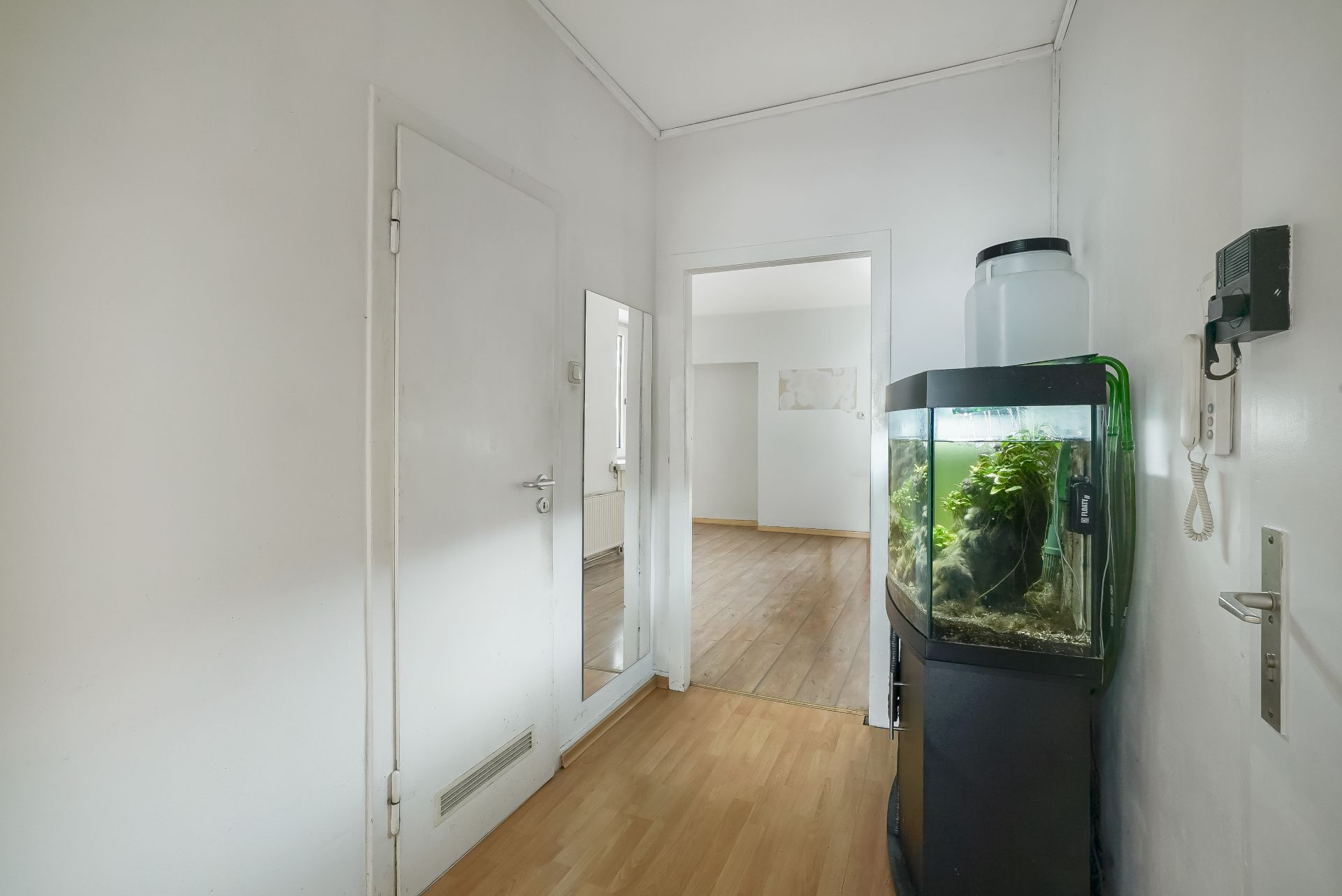 Flur mit Holzboden, Aquarium, Spiegel an der Wand und offener Tür zu einem anderen Raum.