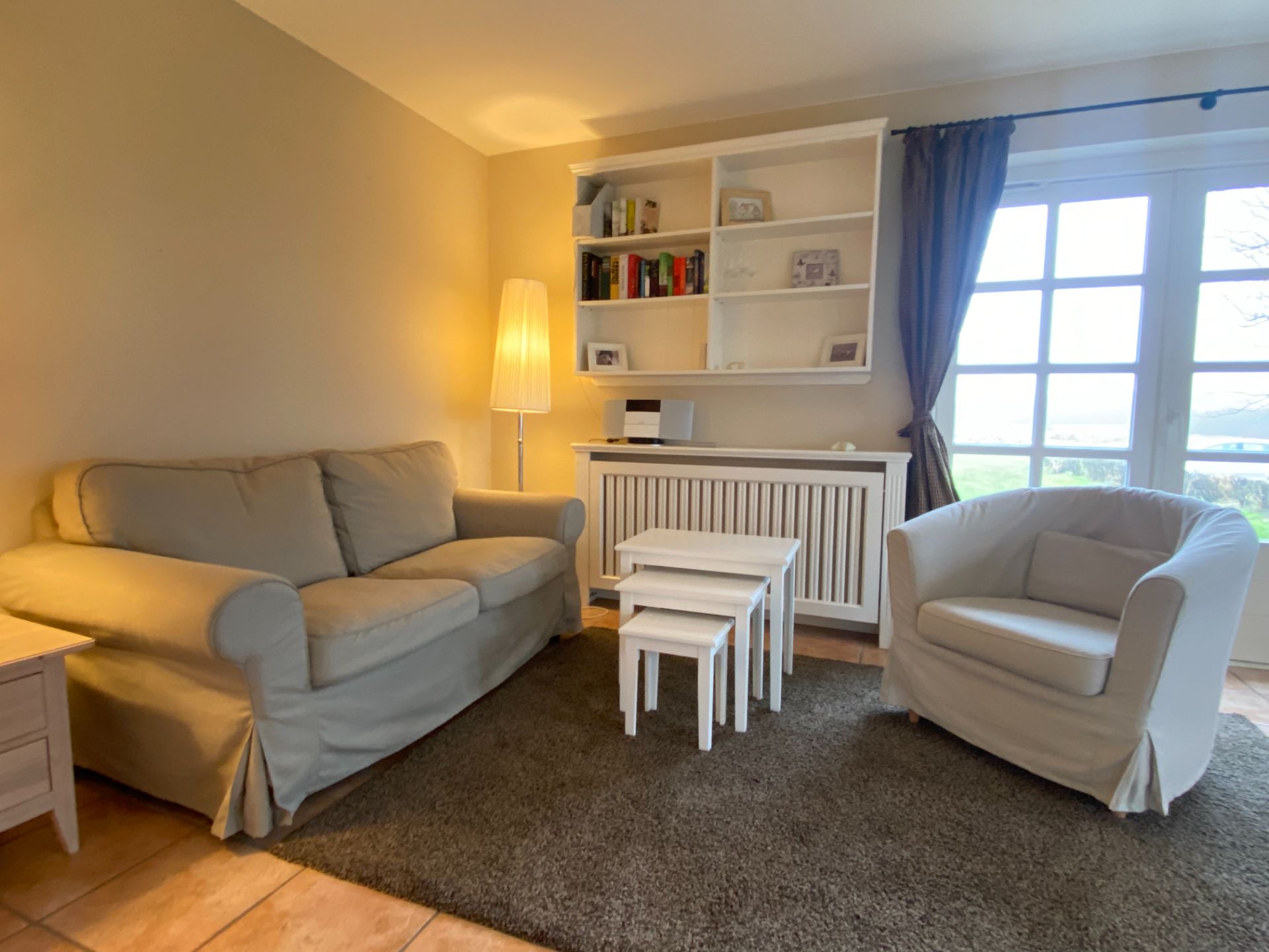Helles Wohnzimmer mit Sofa, Sessel, Nesting-Tischen, Regal, Stehlampe und großem Fenster.