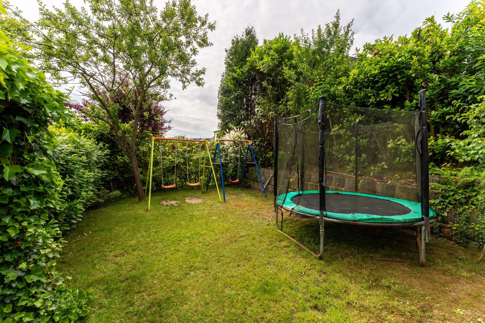 Grüner Garten mit zwei Schaukeln und einem Trampolin, umgeben von Bäumen und Hecken.