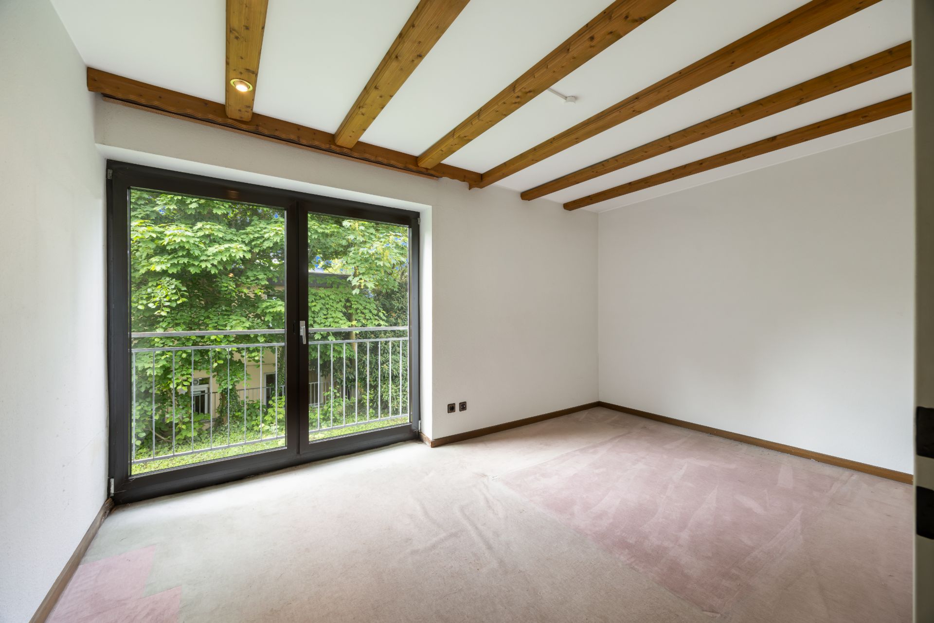 Leeres Zimmer mit weißen Wänden, Holzbalken an der Decke und großer Glastür zum Balkon.