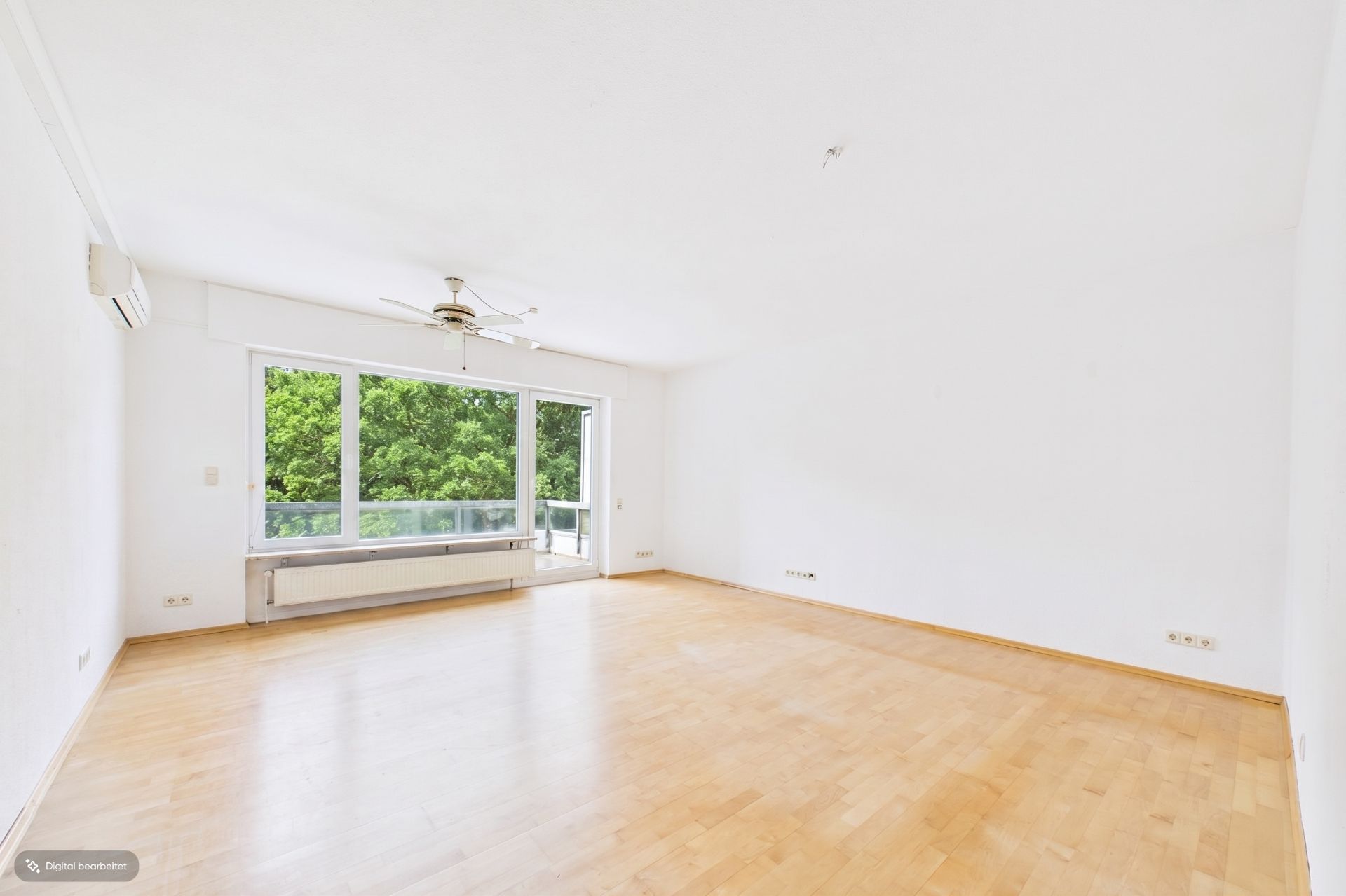 Helles, leeres Zimmer mit Parkettboden, großem Fenster und Deckenventilator.