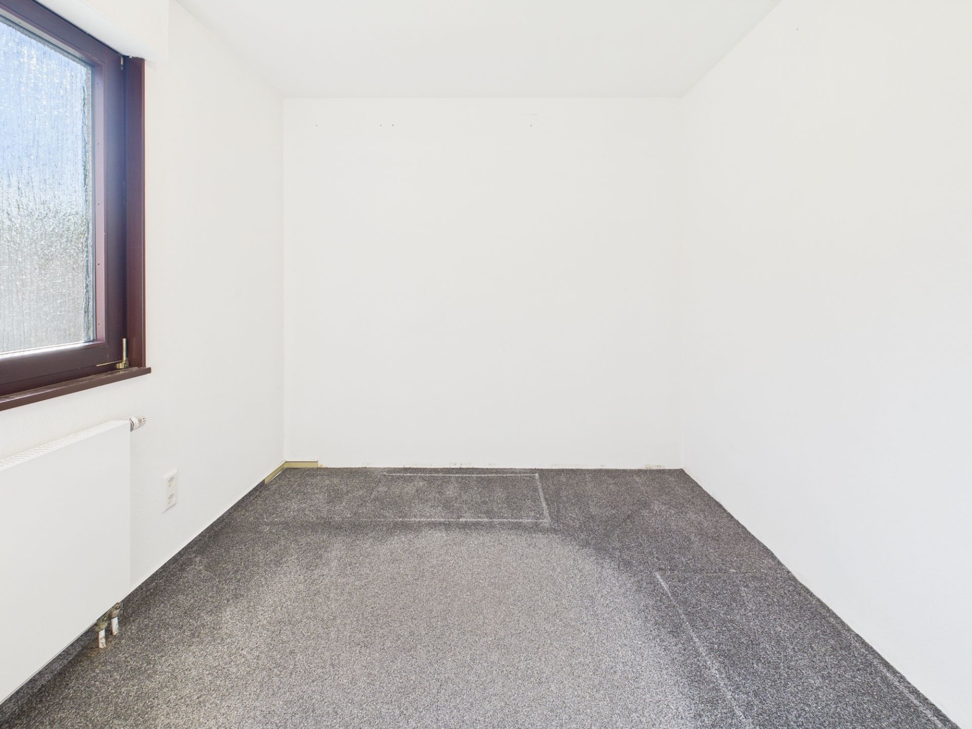 Leerer, kleiner Raum mit grauem Teppich, weißem Wänden und Fenster auf der linken Seite.