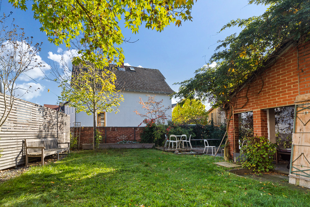 Grüner Garten mit Bank, Bäumen, Rasen und Terrasse mit weißen Stühlen neben einem Backsteingebäude.
