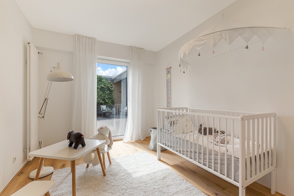 Helles Kinderzimmer mit weißem Babybett, kleinem Tisch, Stuhl, Lampen und Plüschtieren.