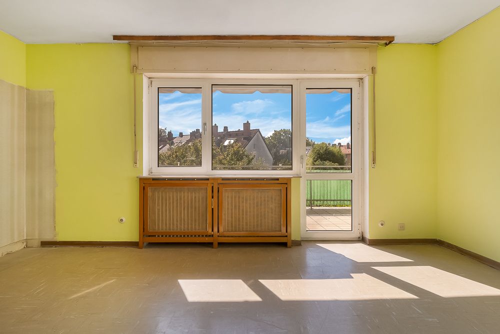 Leeres Zimmer mit gelben Wänden, großem Fenster und Zugang zu einem Balkon.