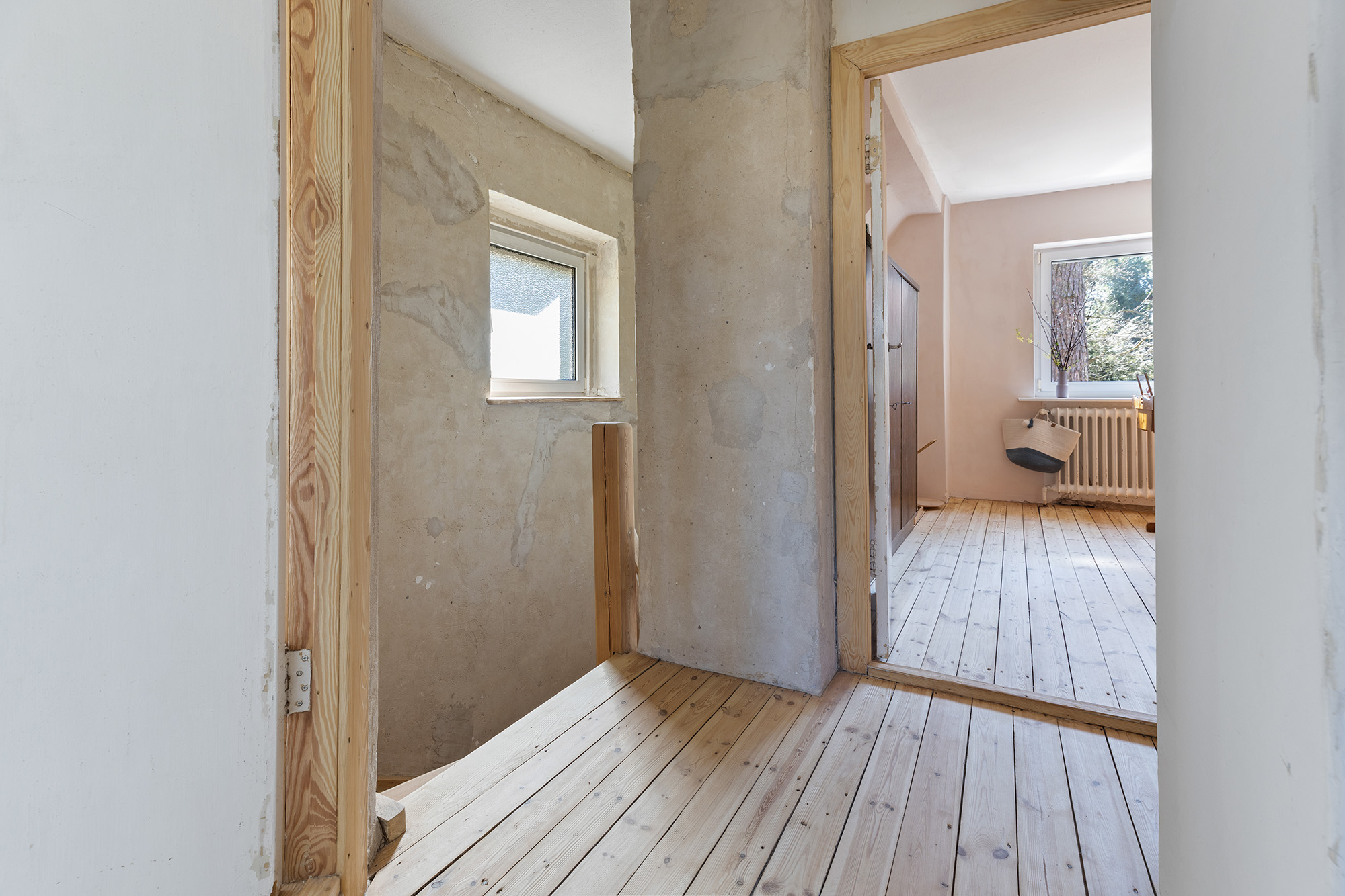 Flur mit Holzboden, roher Wand und Blick in ein helles Zimmer mit Fenster.