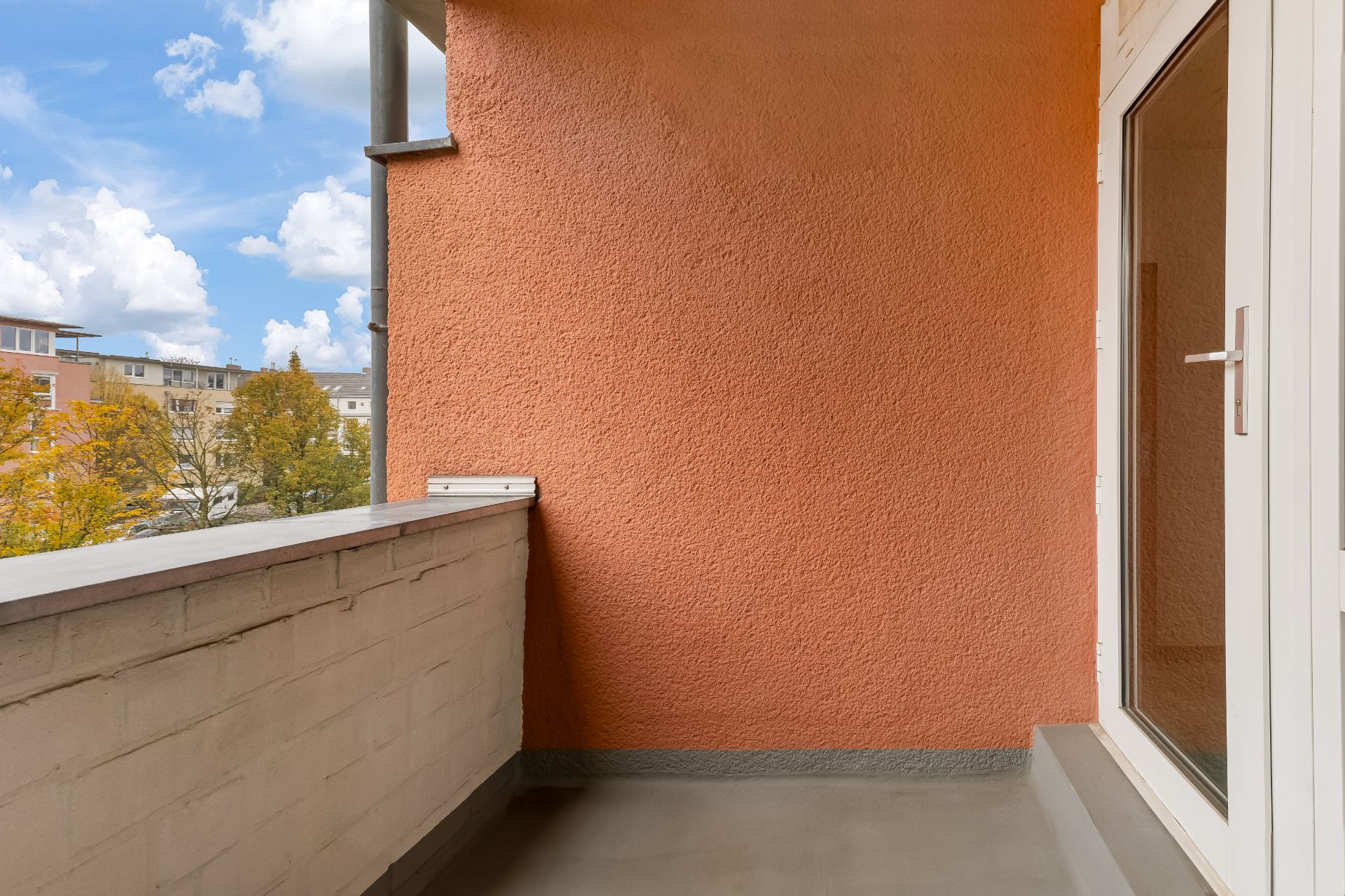 Kleiner Balkon mit oranger Wand und Glastür, Blick auf Bäume und Nachbargebäude.