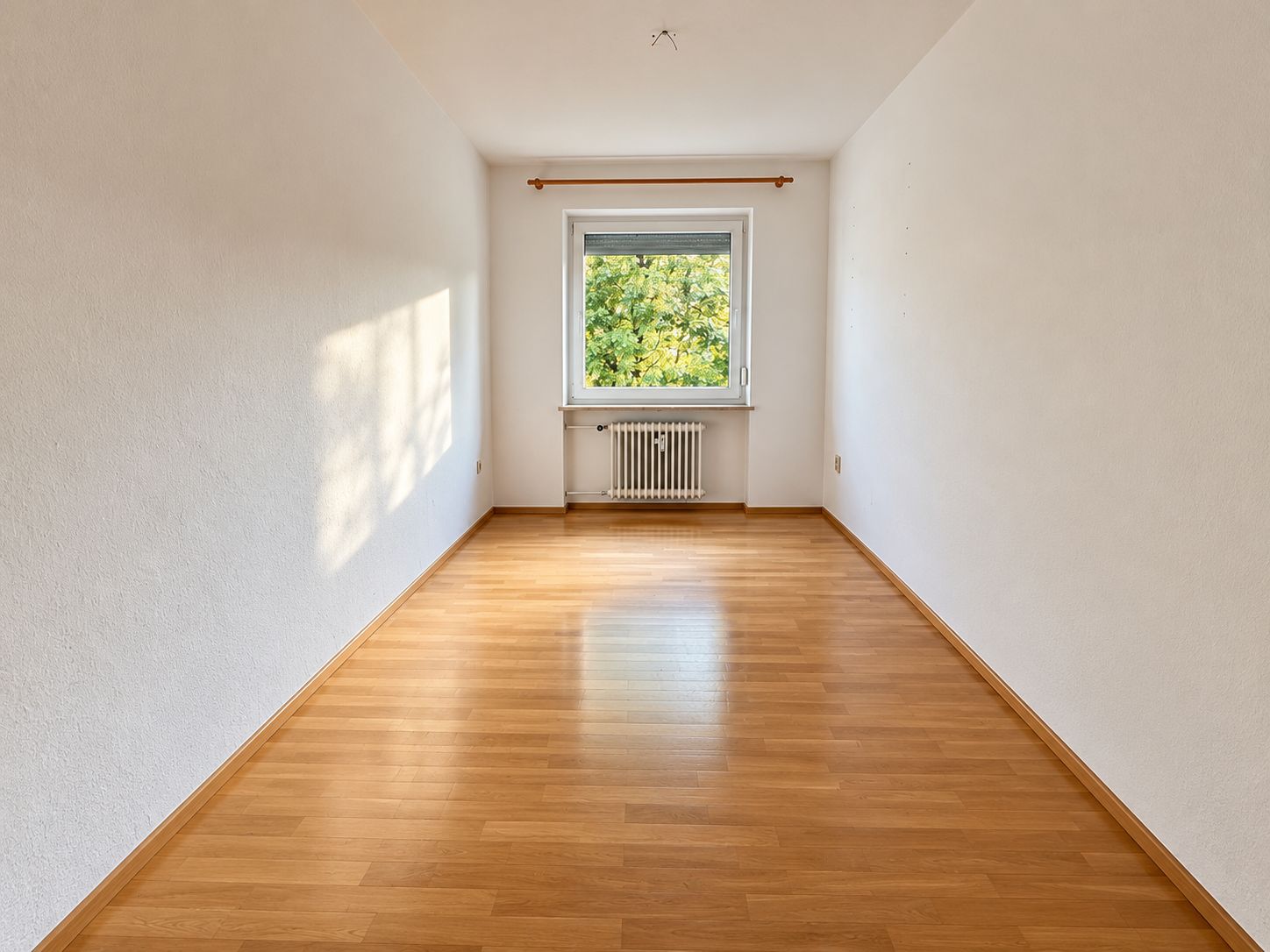 Leerer, heller Raum mit Parkettboden, weißem Fenster und Heizung unter dem Fenster.