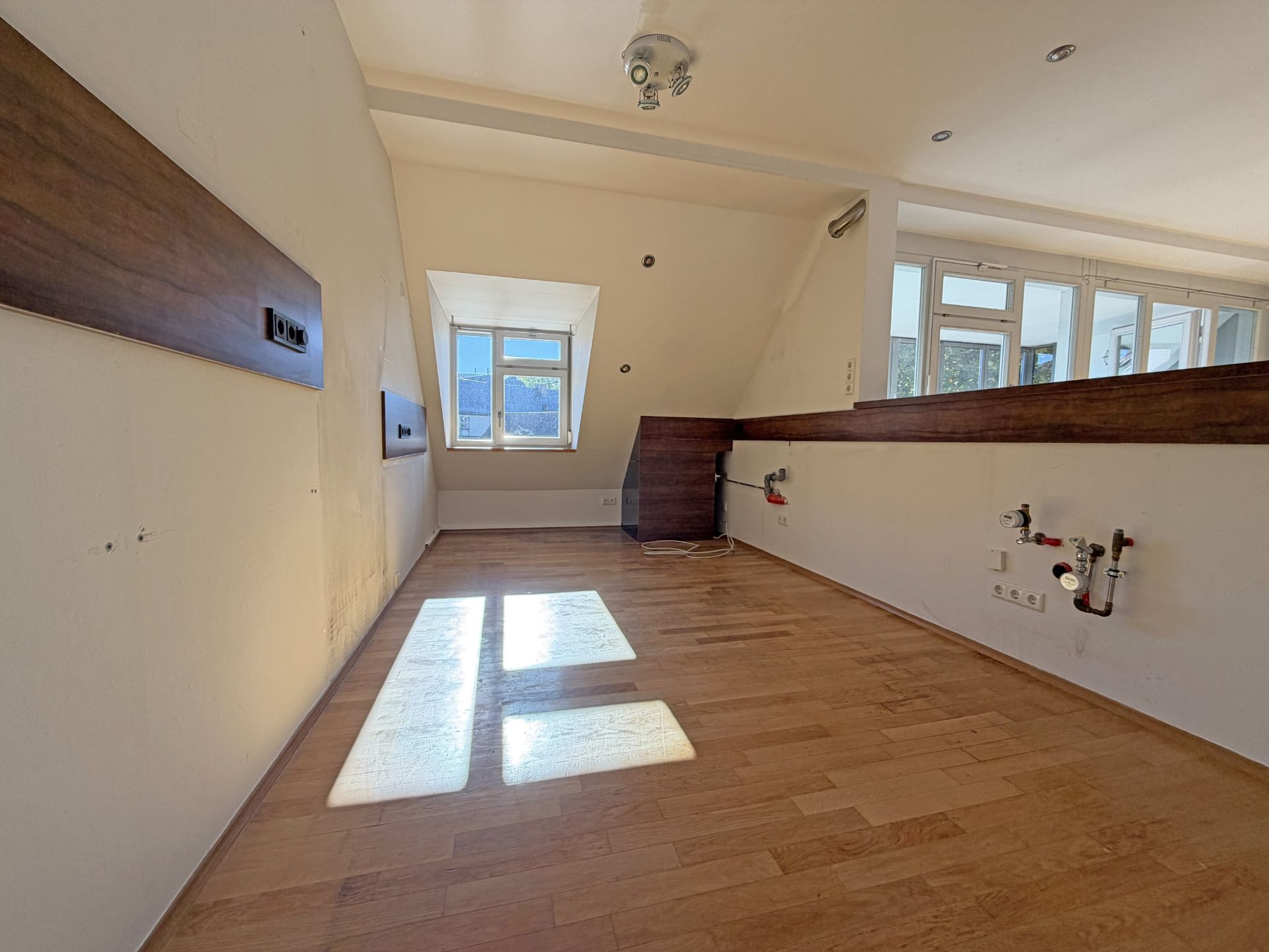 Leerer Raum mit Holzboden, Dachfenster und Anschlüssen an der Wand.