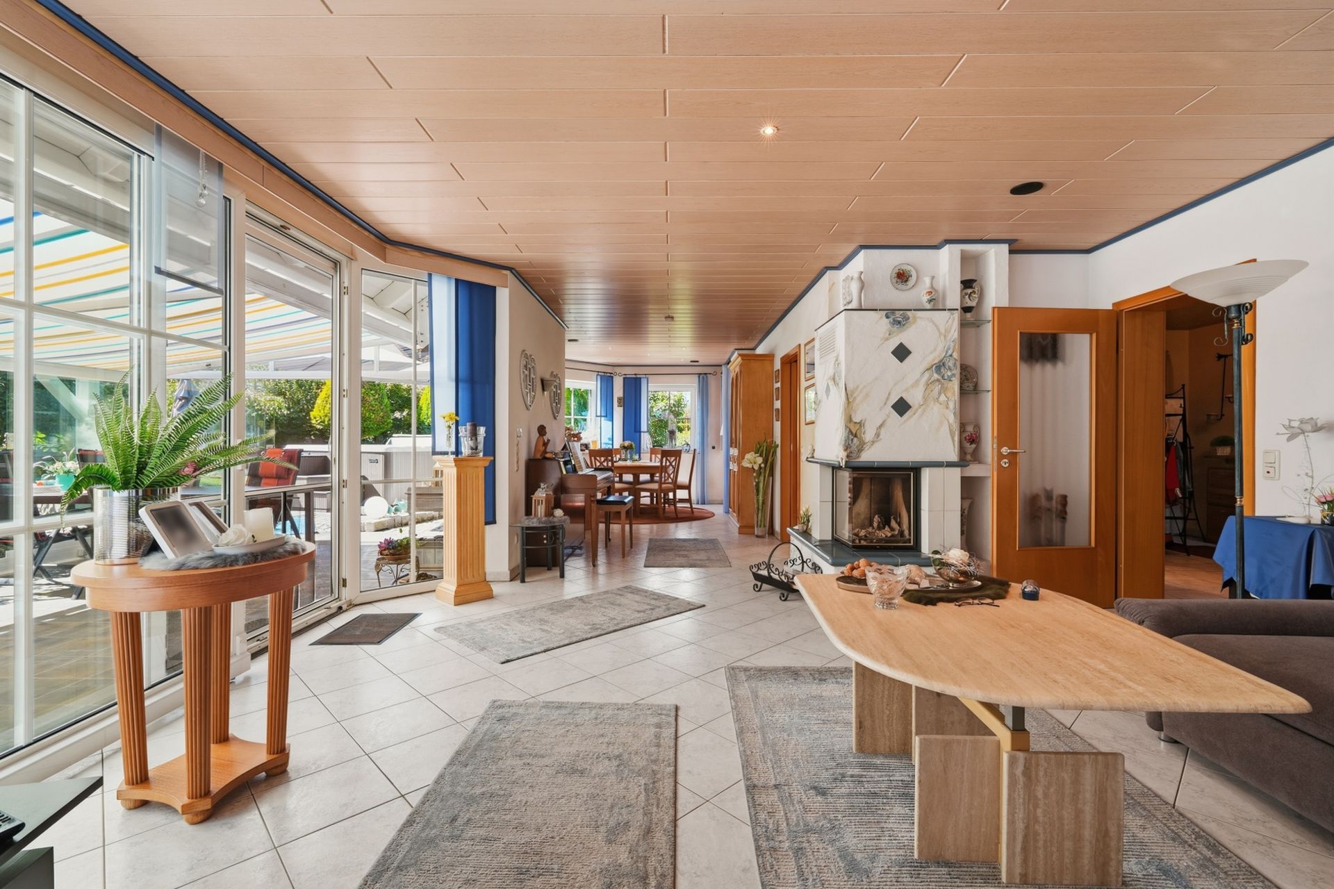 Helles Wohnzimmer mit Holztisch, Kamin, Teppichen und Zugang zur Terrasse durch große Glasfenster.