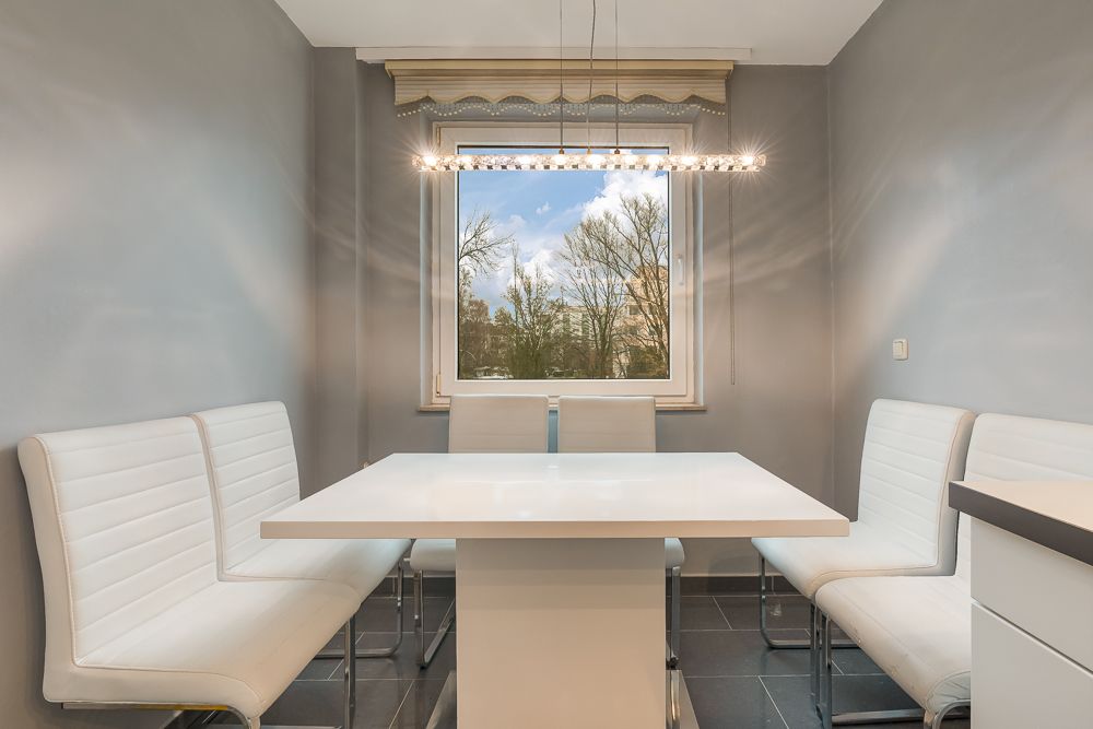 Moderner Essbereich mit weißem Tisch und weißen Stühlen vor einem Fenster mit Blick nach draußen.