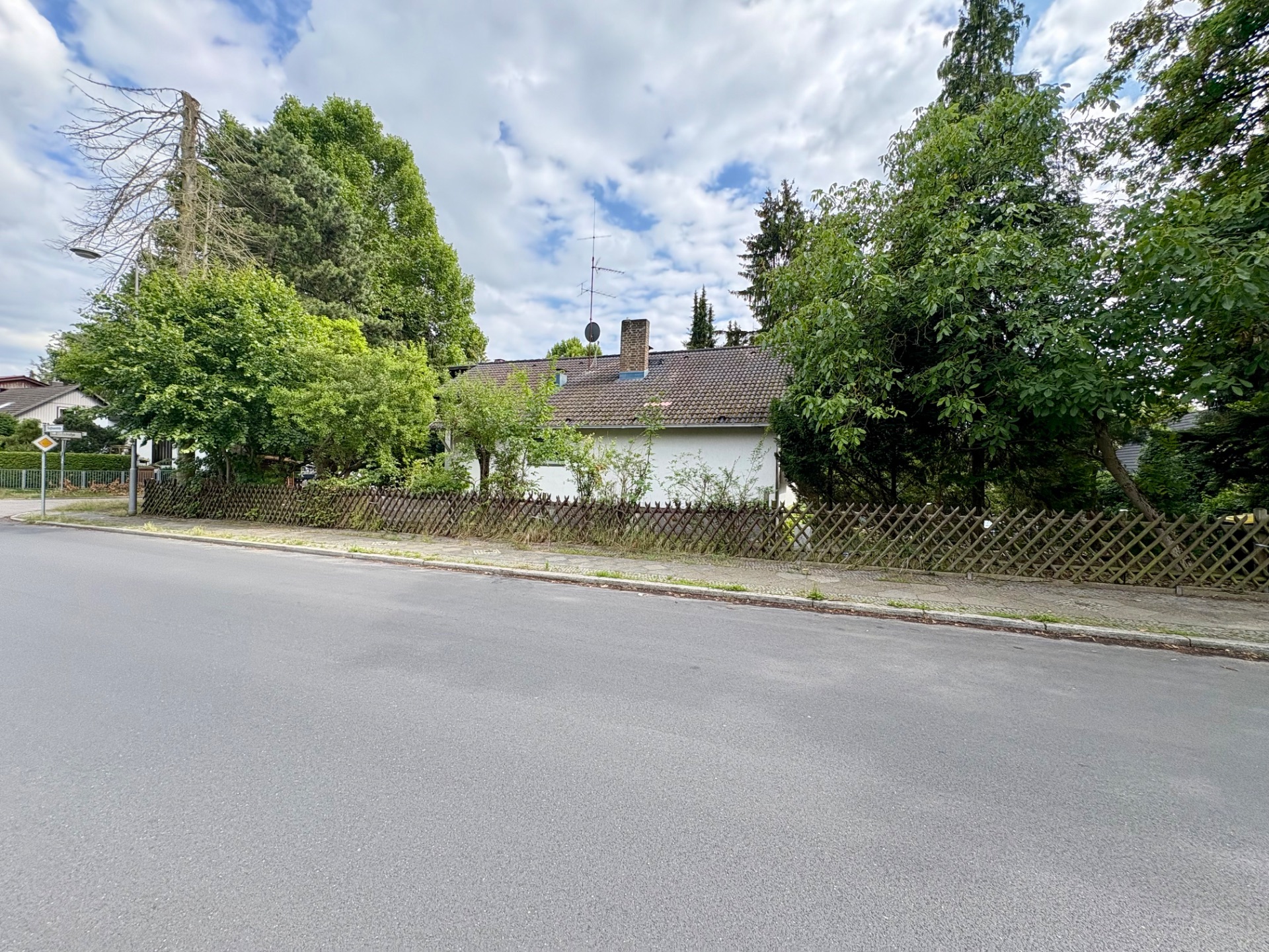 Einfamilienhaus mit schrägem Dach, umgeben von Bäumen und Holzzaun an einer ruhigen Straße.