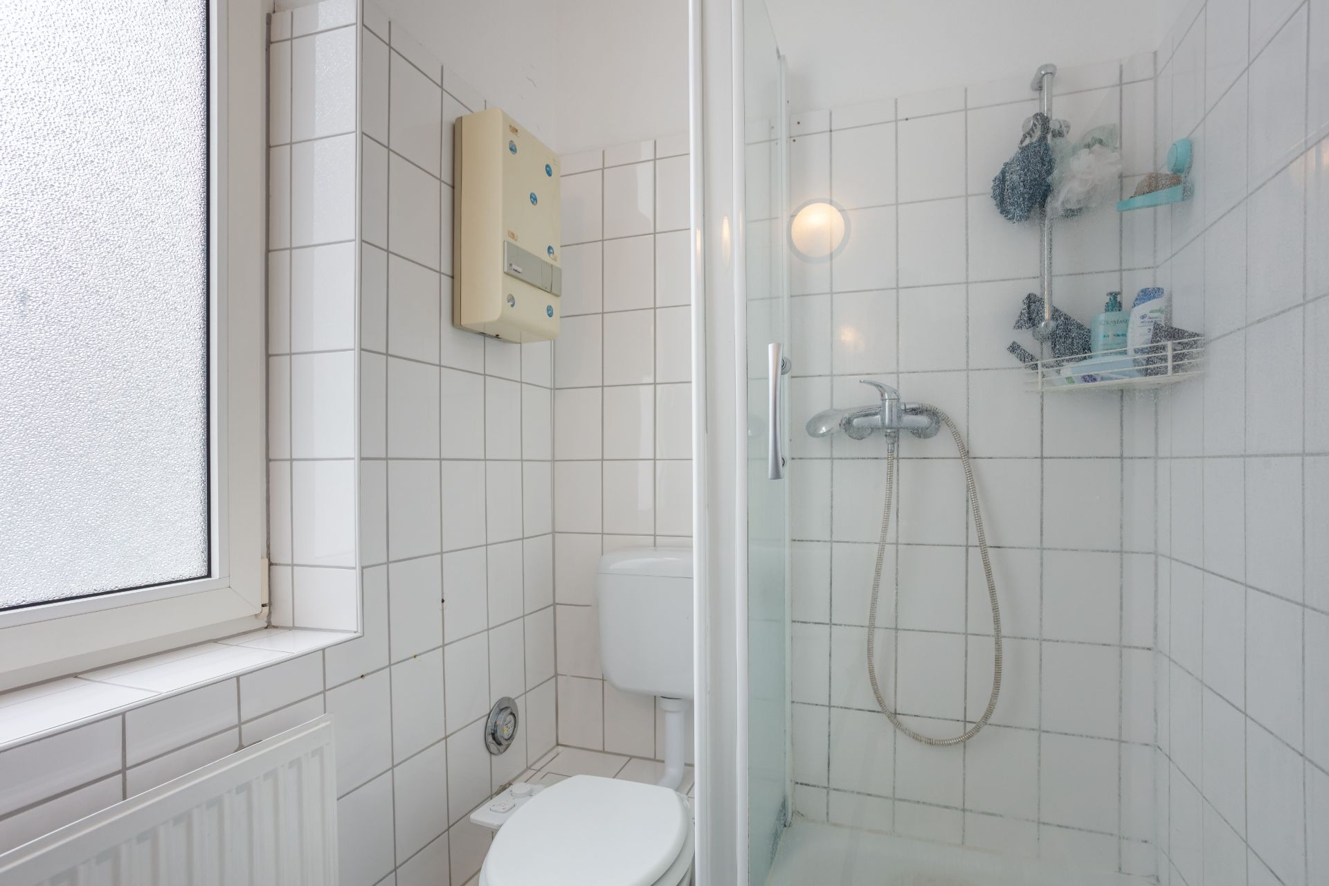 Kleines Badezimmer mit Dusche, Toilette und Fenster. Weiße Fliesen an den Wänden.