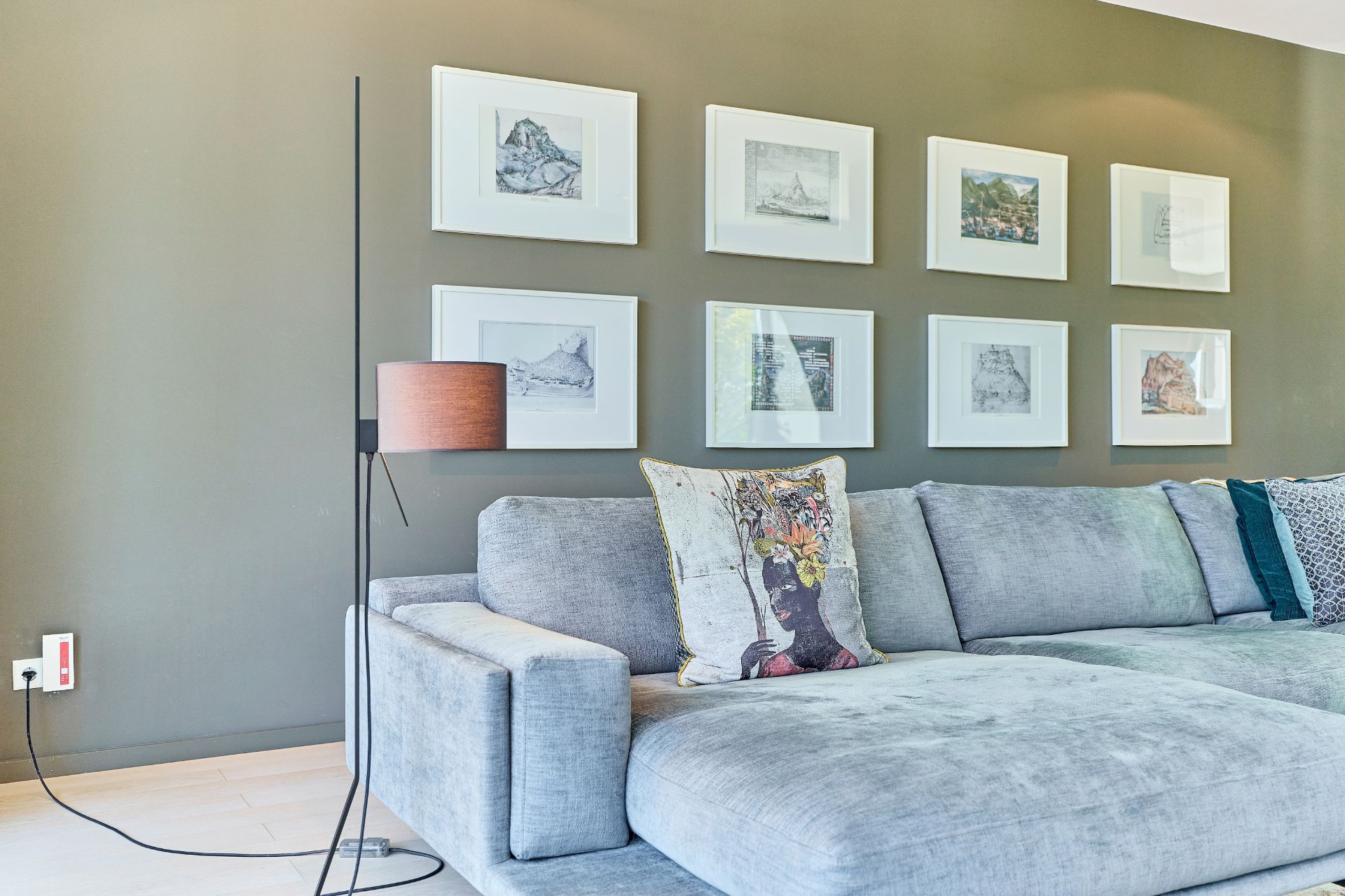 Graues Sofa mit bunten Kissen vor grüner Wand mit acht gerahmten Bildern.