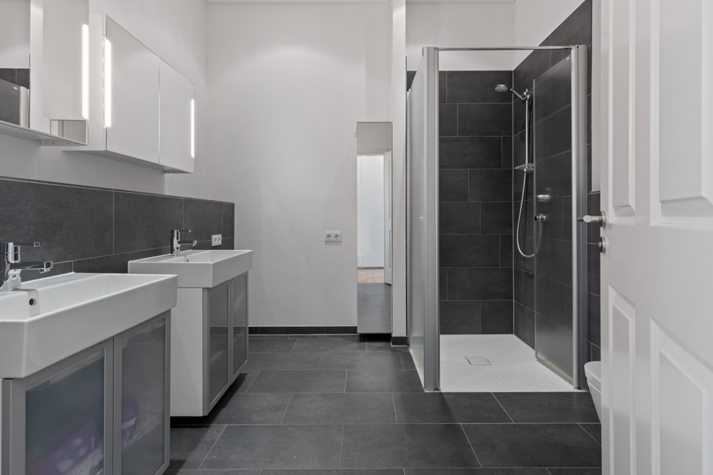 Modernes Badezimmer mit zwei Waschbecken und einer Dusche mit Glaswand und dunklen Fliesen.